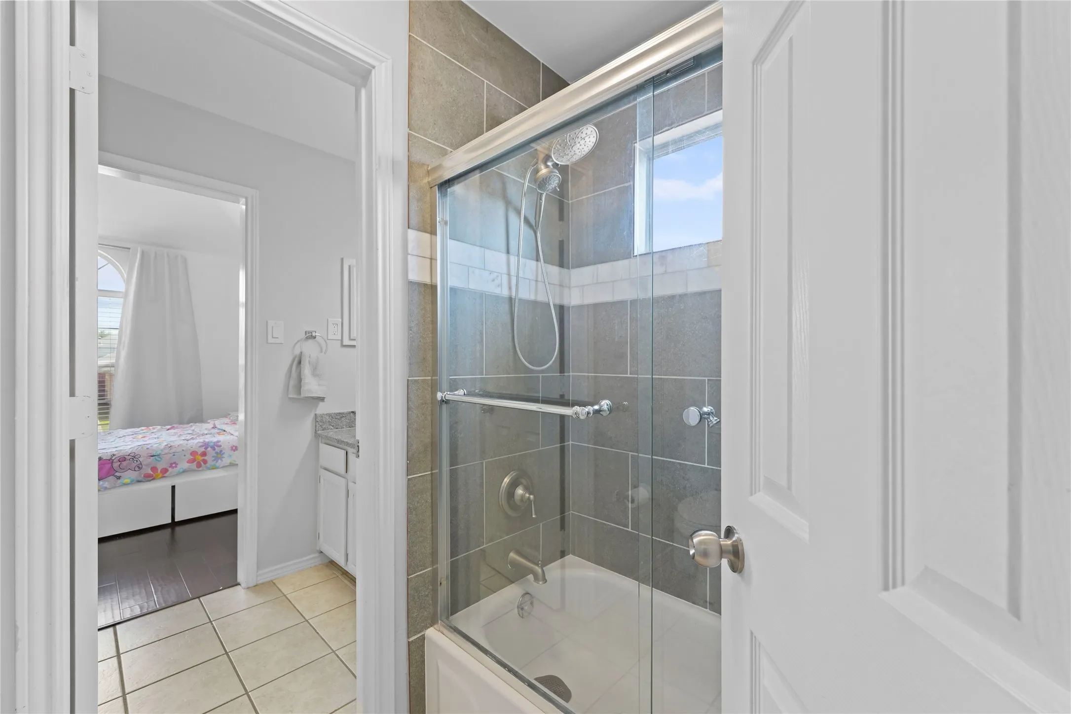 Shower-tub combination featuring a glass enclosure and gray tile surround with decorative accent band, complemented by a white vanity with a stone countertop and an arched window in the adjacent room