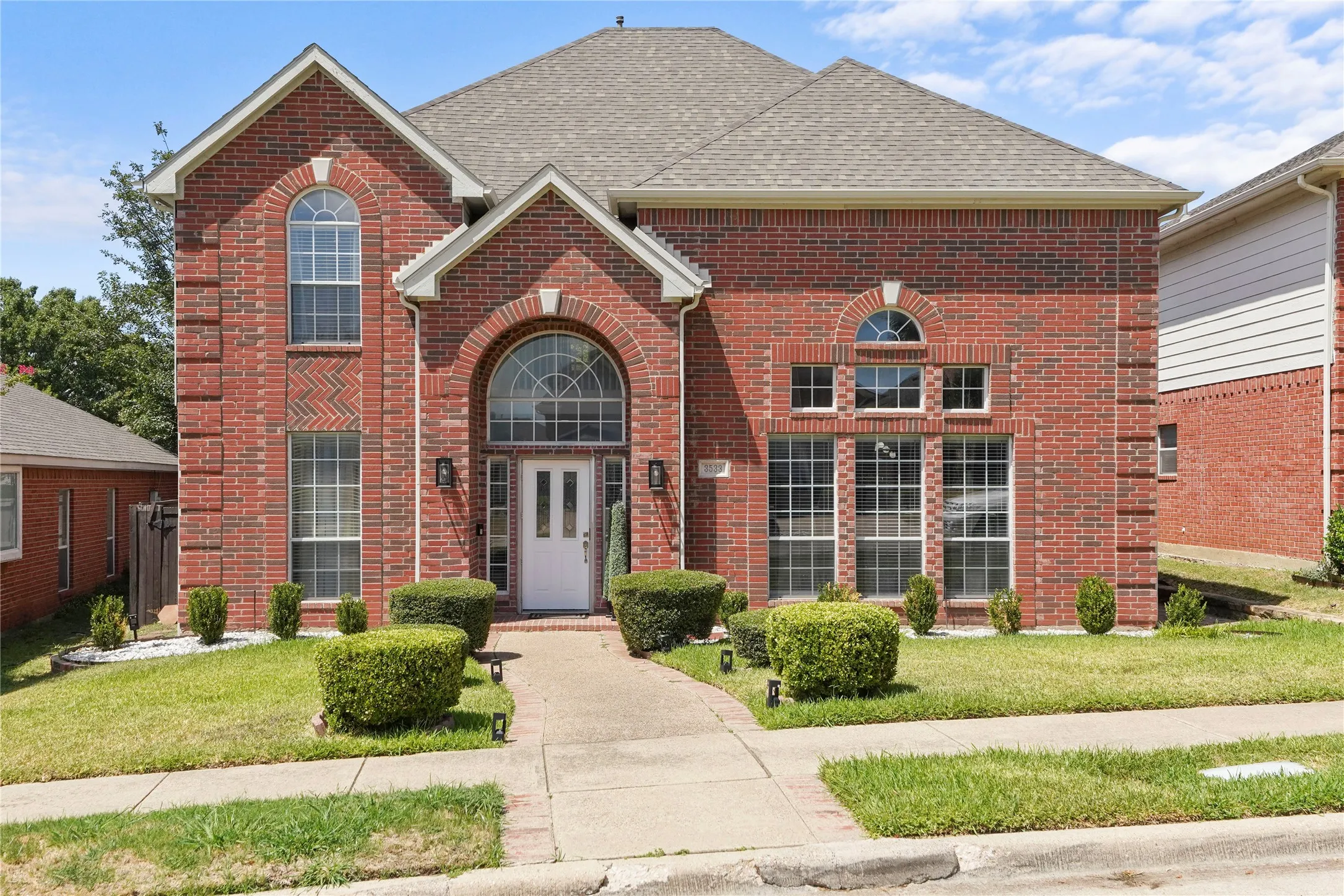 Red brick exterior with arched windows, a prominent arched entry, white front door, landscaped front yard with trimmed hedges, and a concrete walkway leading to the entrance
