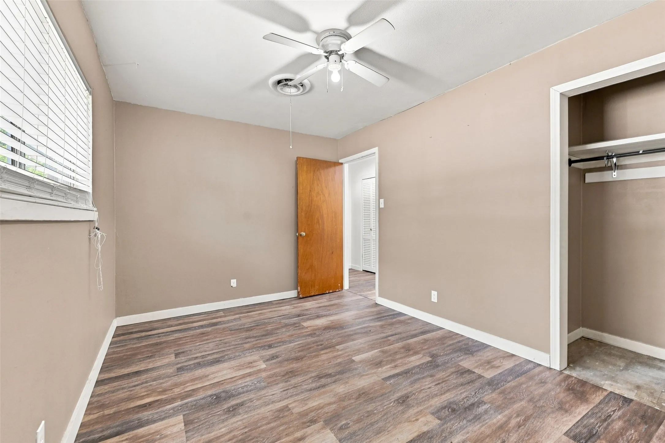 Wood-finish flooring, ceiling fan with light fixture, window with blinds, and built-in closet with shelving and hanging rod