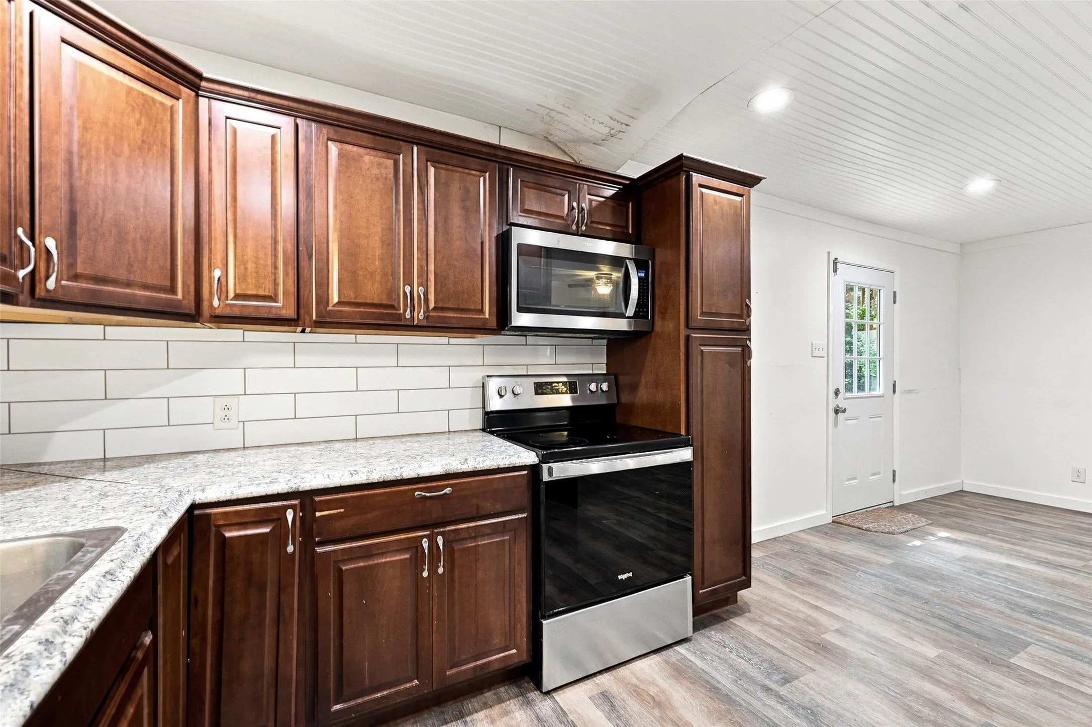 Dark wood cabinetry, white subway tile backsplash, light stone-finish countertops, stainless steel appliances, and wood-finish flooring