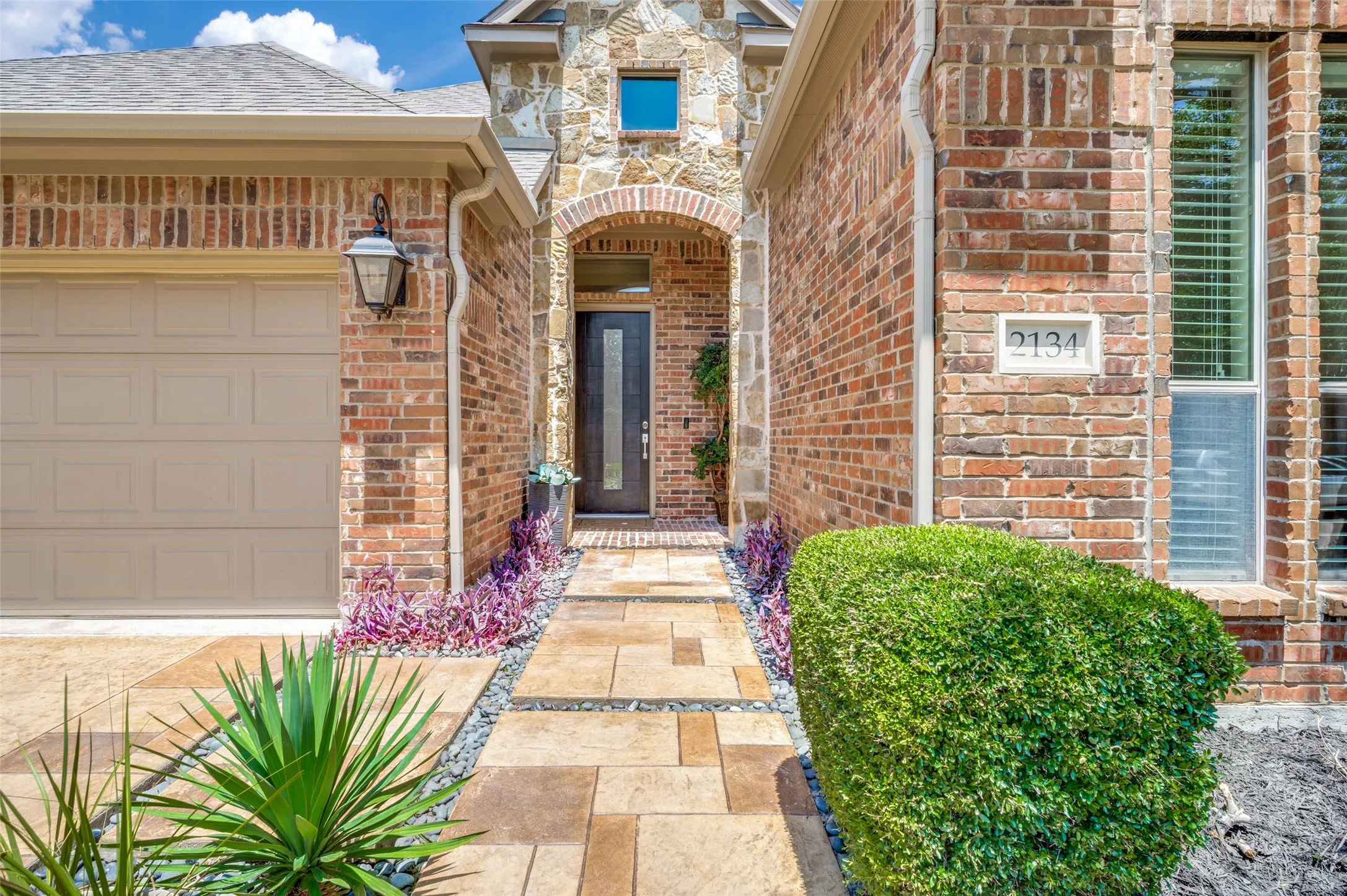 A charming approach welcomes you home with an upgraded custom front door framed by an elegant stone entry and covered front porch. The custom stone walkway creates a warm first impression, while lush professional landscaping, manicured shrubs, decorative rock accents, and mature trees enhance the home's exceptional curb appeal.