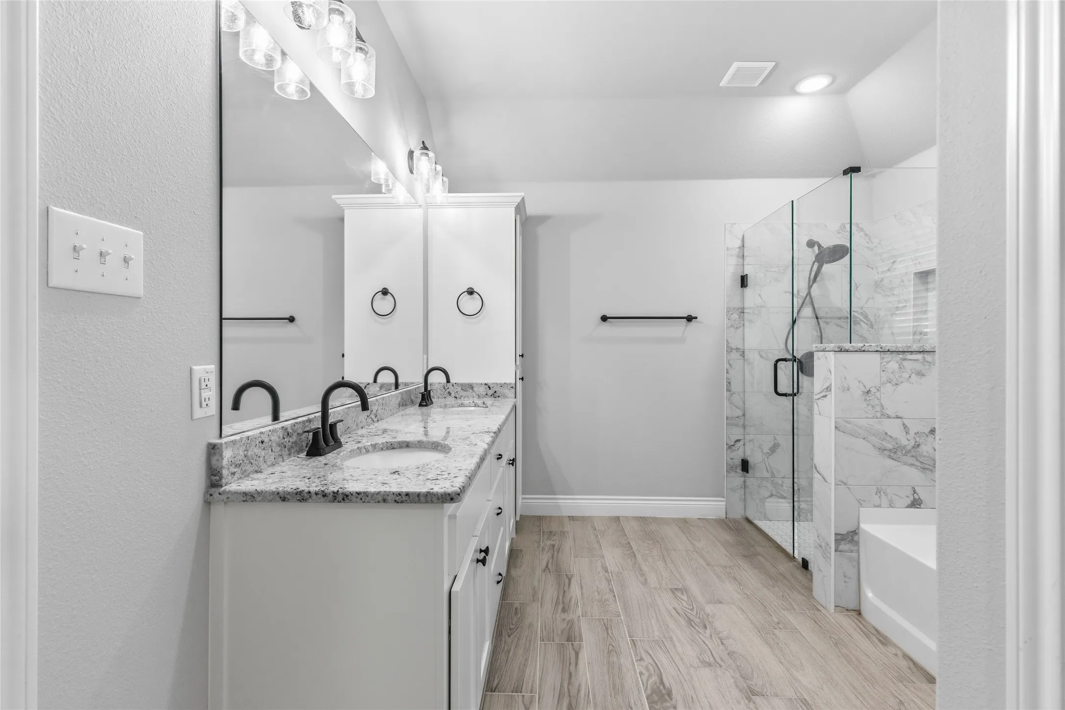 Spacious bathroom featuring a dual vanity with granite countertops, built-in storage cabinets, wood-finish flooring, and a glass-enclosed shower with marble-look tile surround