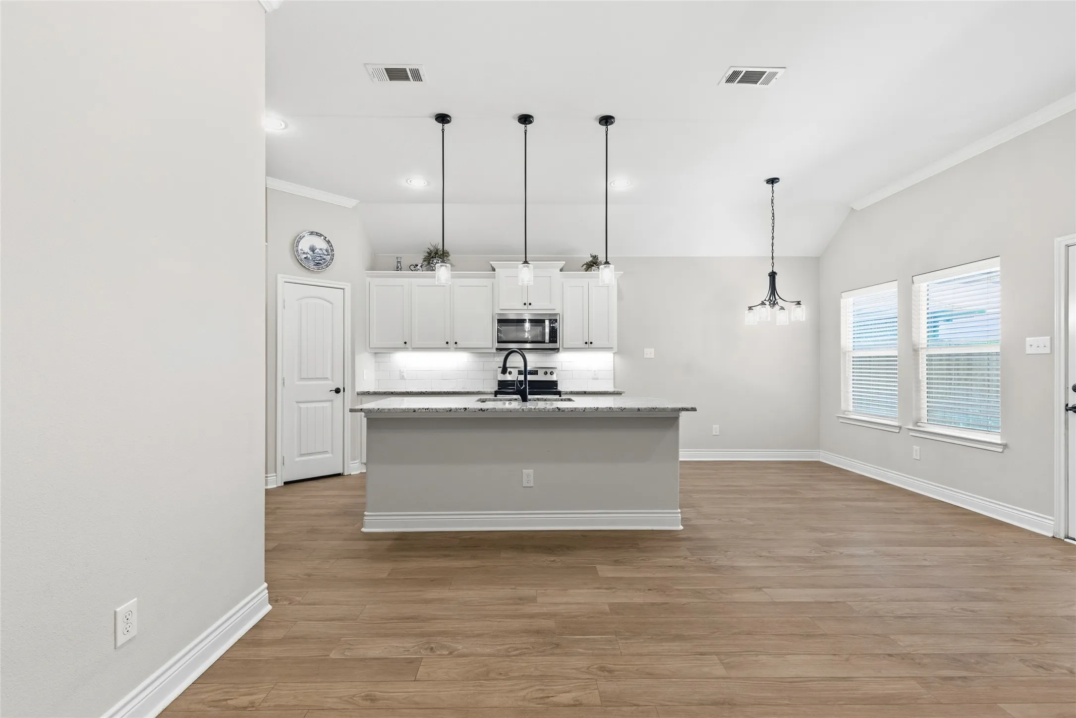 Open-concept living space featuring wood-finish flooring, a kitchen island with pendant lighting, white cabinetry, integrated stainless steel appliances, and vaulted ceilings