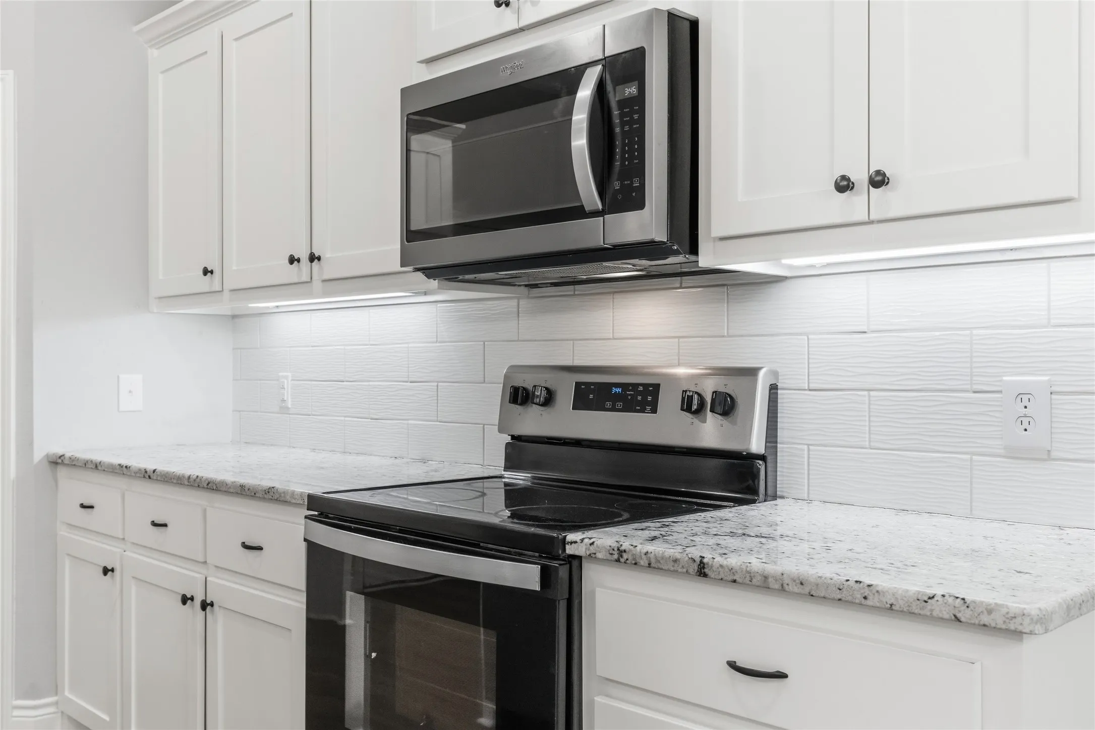 Kitchen featuring white cabinetry with dark hardware, granite countertops, white textured subway tile backsplash, stainless steel microwave, and a black electric range