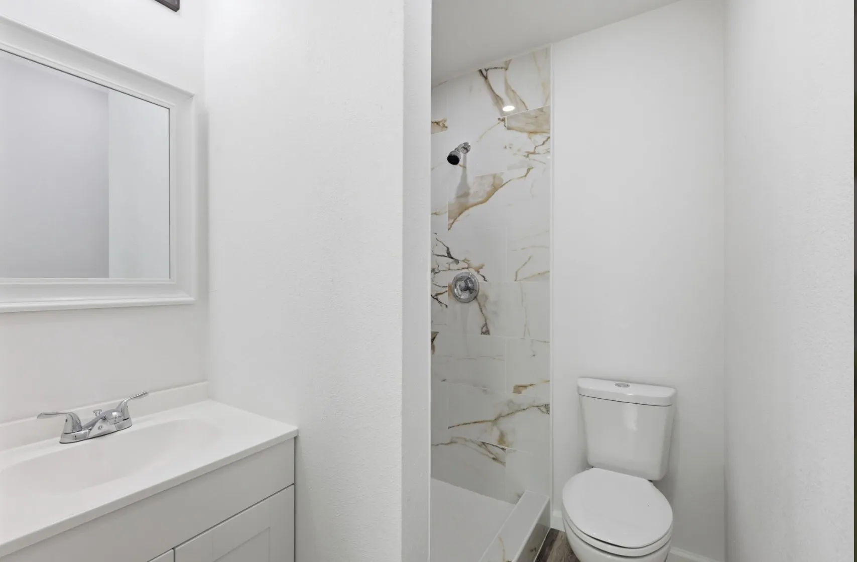 Compact bathroom featuring a white vanity with integrated sink, framed mirror, and a shower with white and gold veined tile surround