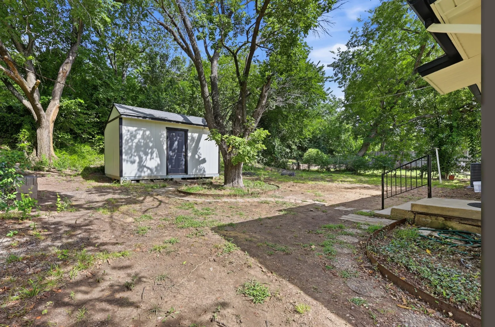 Spacious backyard featuring a white storage shed with dark trim, mature trees, a concrete patio with black metal railing, and stepping stones