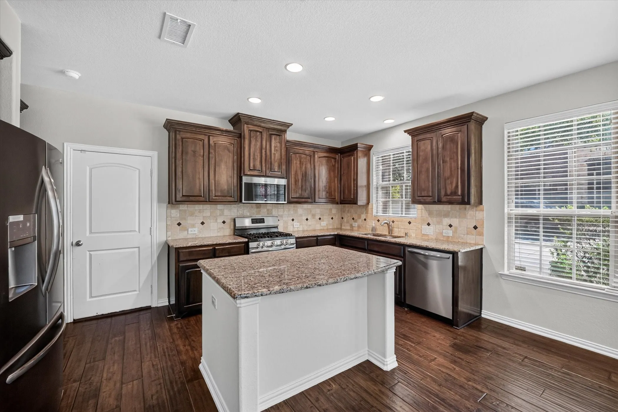 Kitchen featuring a central island with a granite countertop, wood-finish flooring, dark wood cabinetry, stainless steel appliances, and tile backsplash