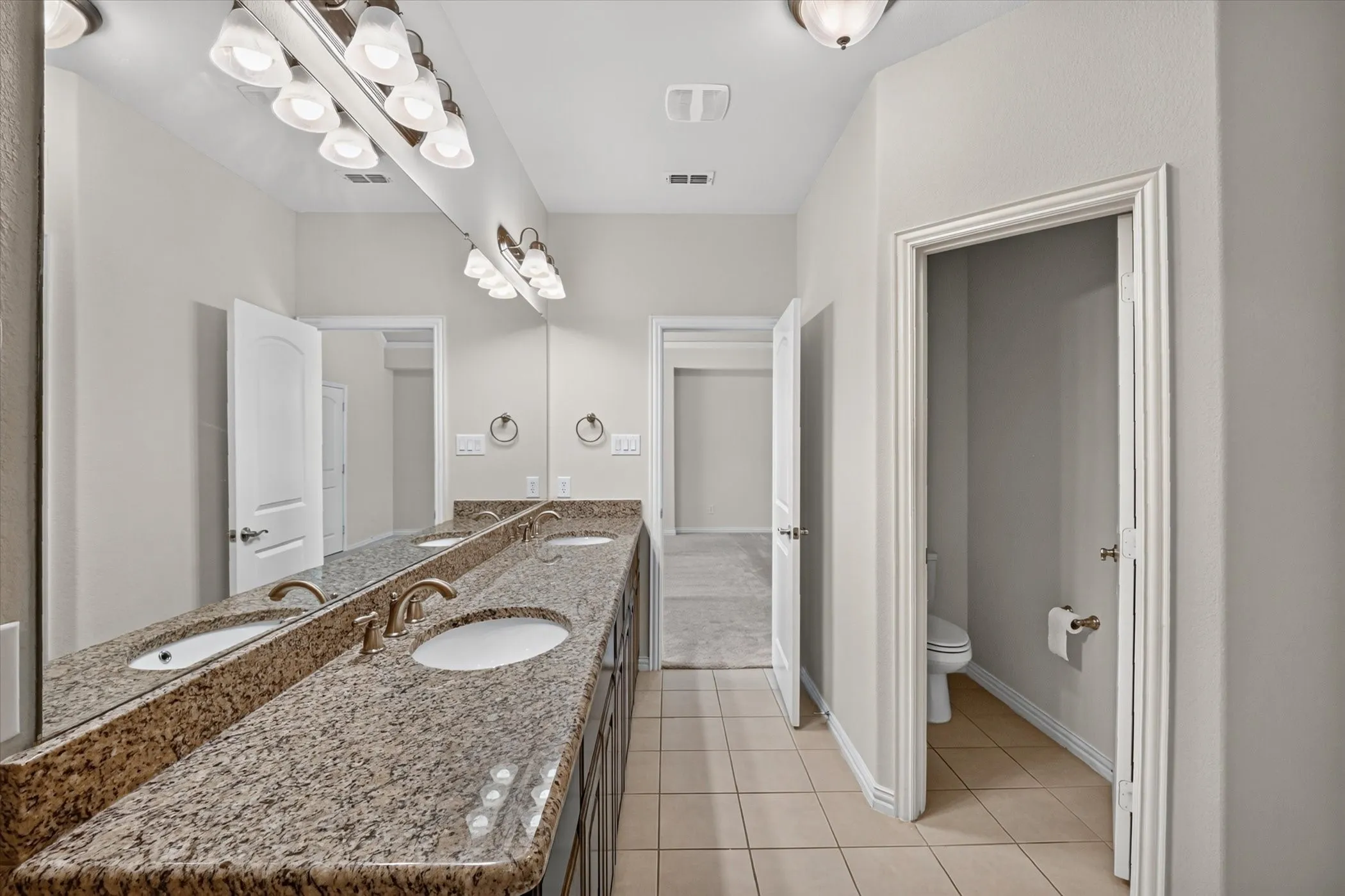 Double vanity featuring a granite-style countertop with dual undermount sinks, bronze-finish fixtures, and full-width mirror with overhead sconce lighting