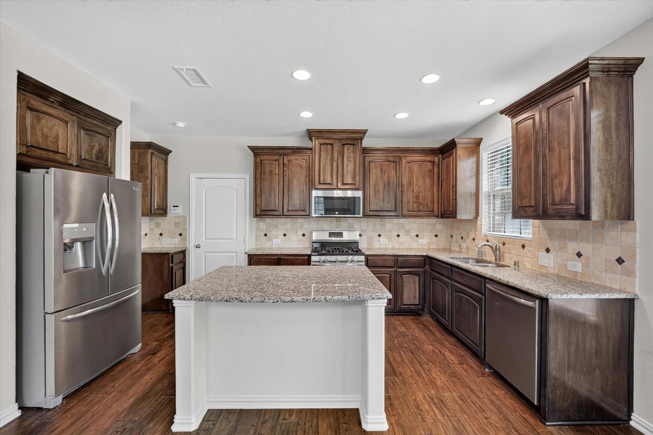 Kitchen featuring an island with a light-toned countertop, wood-finish flooring, dark wood cabinetry, stainless steel appliances, and recessed lighting