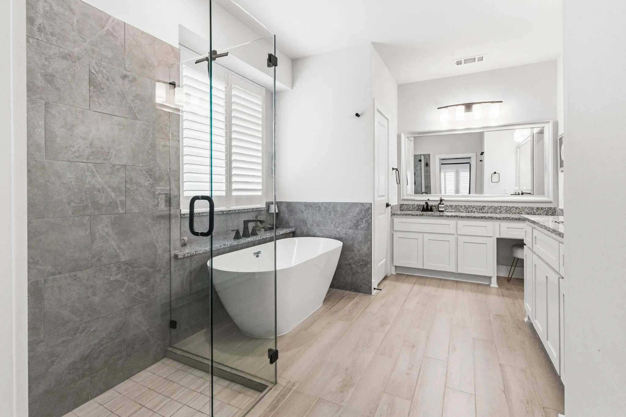 Modern bathroom with a spacious glass-enclosed shower featuring large format gray tile, a freestanding soaking tub, wood-finish flooring, a double vanity with granite countertops, and white cabinetry