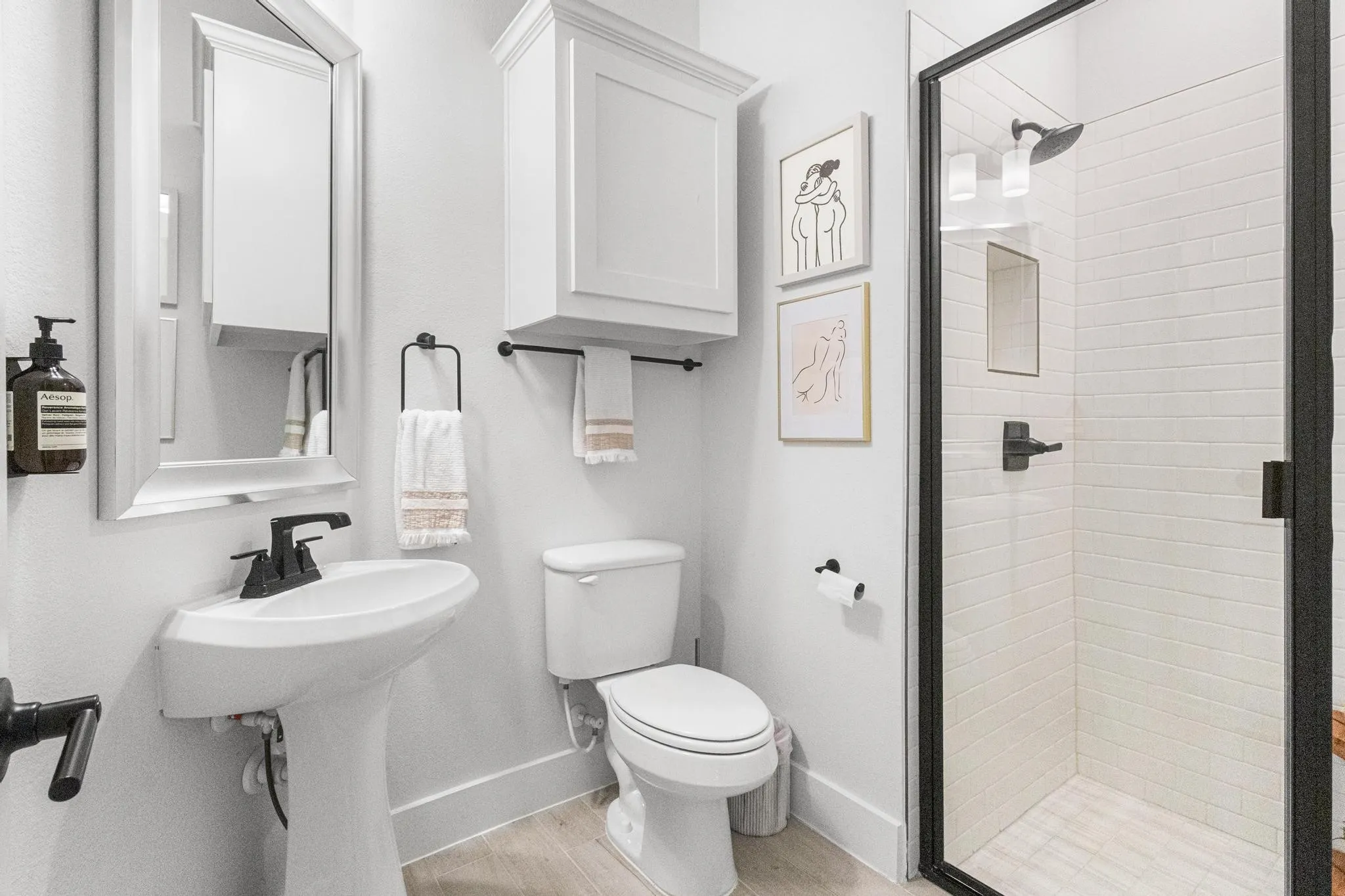 Modern bathroom featuring a glass-enclosed shower with subway tile surround, a pedestal sink with a matte black faucet, and an overhead storage cabinet