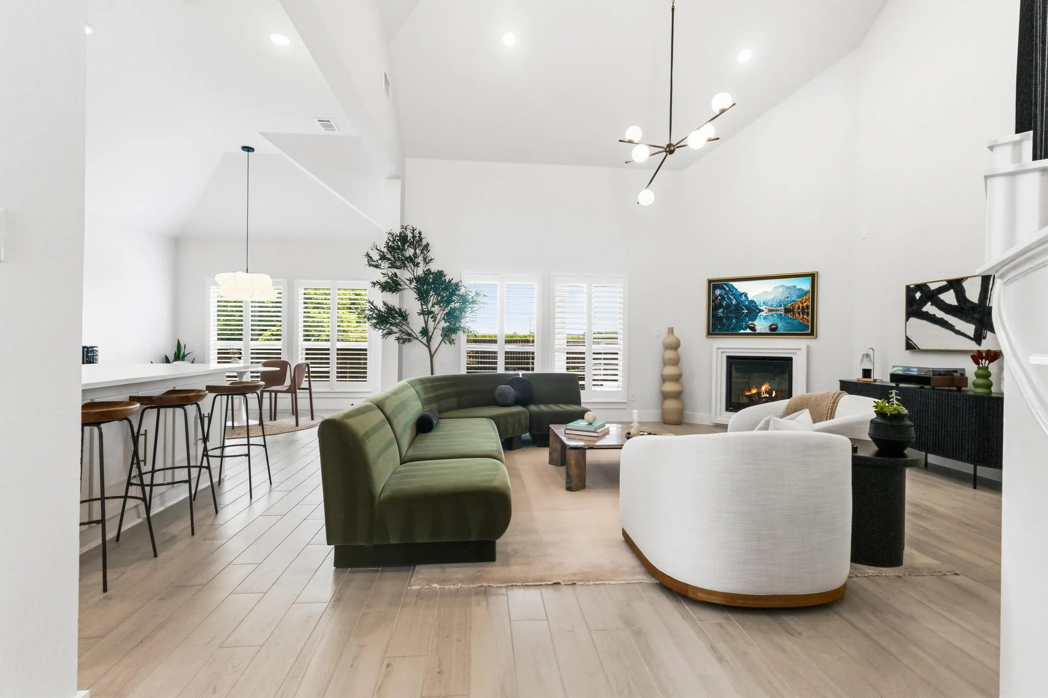 Spacious living area featuring high vaulted ceilings, light wood-finish flooring, a fireplace with a white mantel, and white plantation shutters on multiple windows