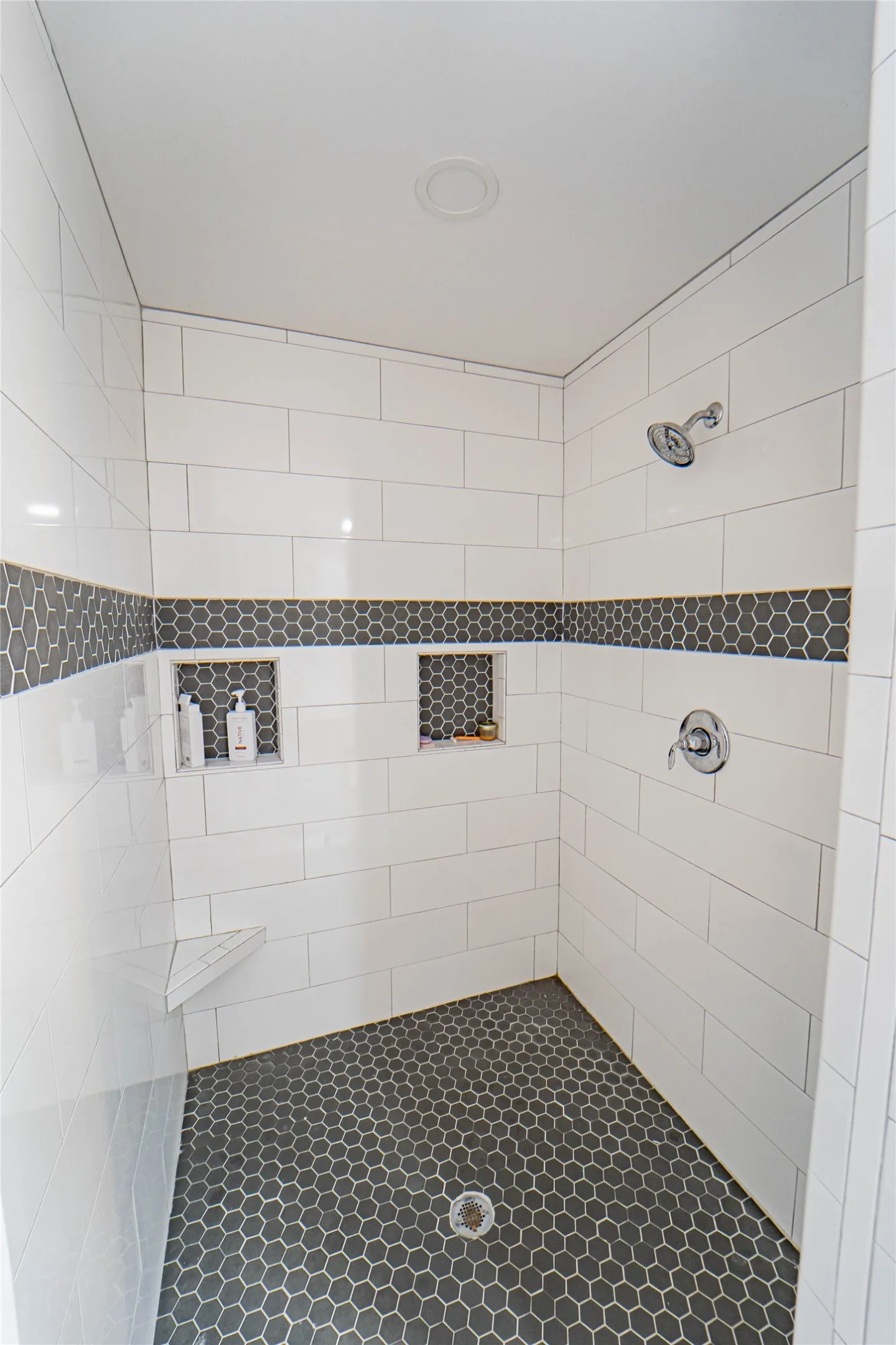 Shower enclosure featuring white rectangular wall tiles, a dark gray hexagonal floor, and a coordinating accent tile band