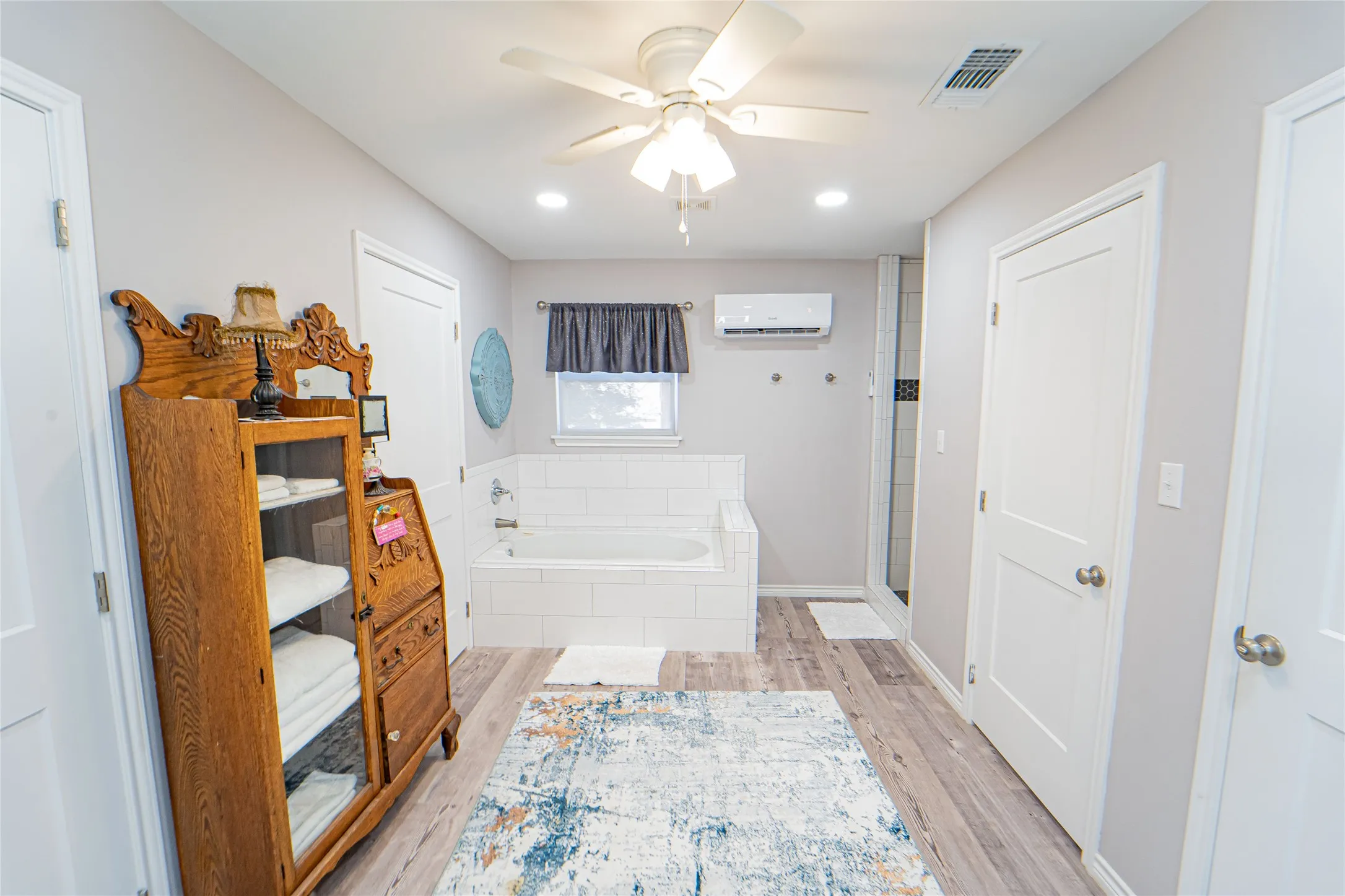 Spacious bathroom featuring a built-in soaking tub with white subway tile surround, separate glass-enclosed shower, wood-finish flooring, a ceiling fan with integrated lighting, and recessed lighting