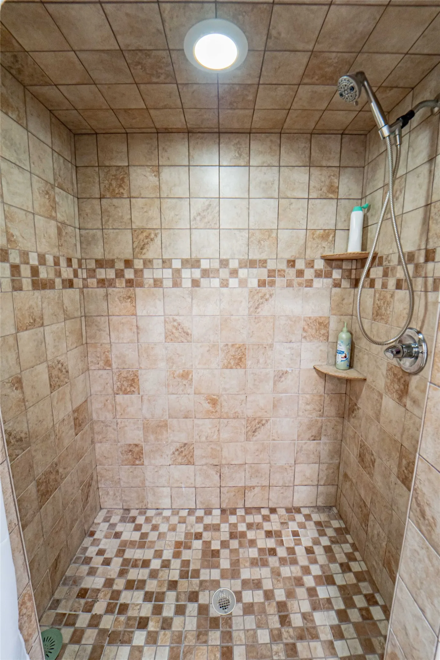Tiled shower with a mosaic floor, decorative accent tile band, recessed ceiling light, and built-in corner shelves