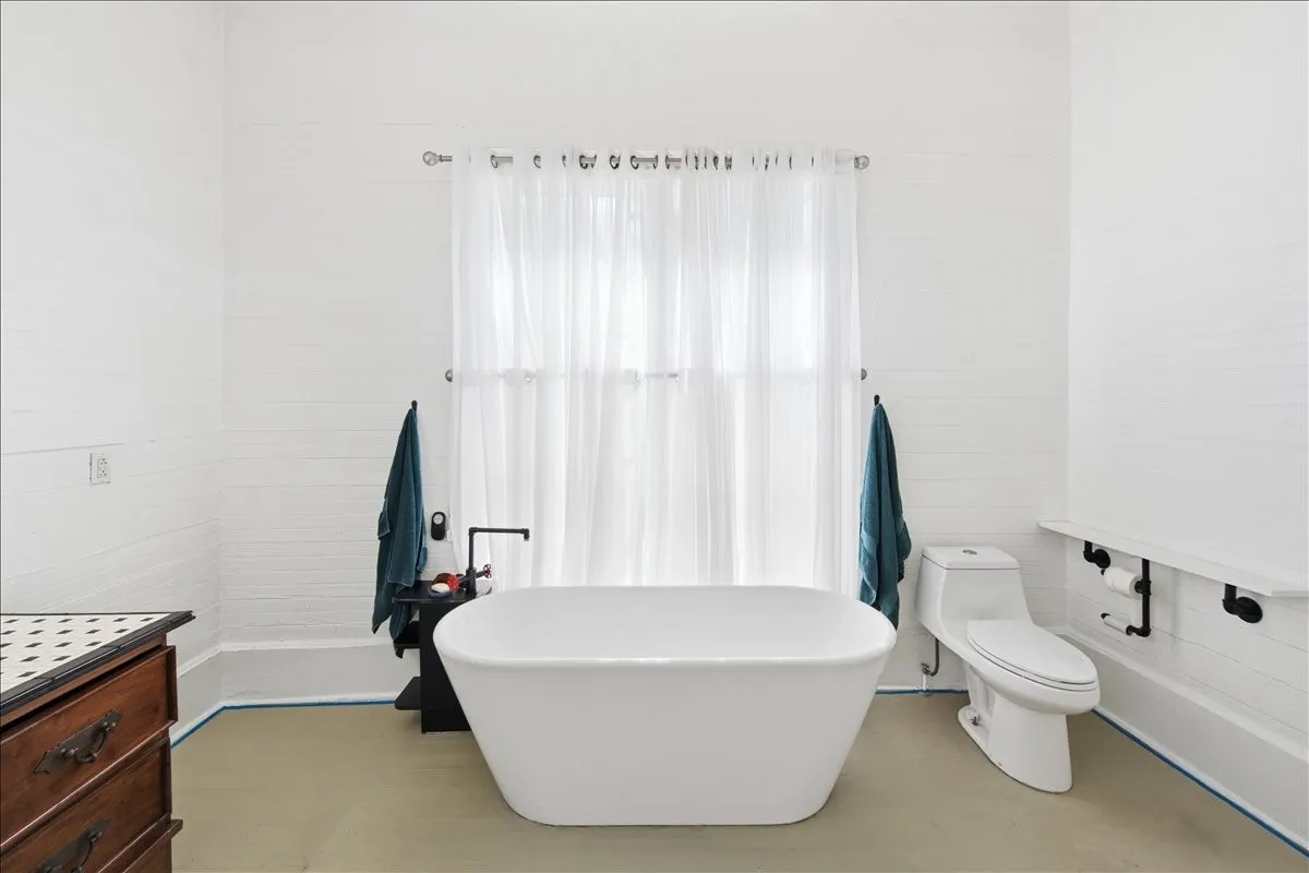 Spacious bathroom featuring a freestanding soaking tub, large window with sheer curtains, and a dark wood vanity.