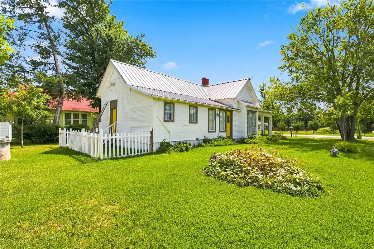 Charming white cottage exterior framed by a white picket fence, green grass, and lush surrounding trees.