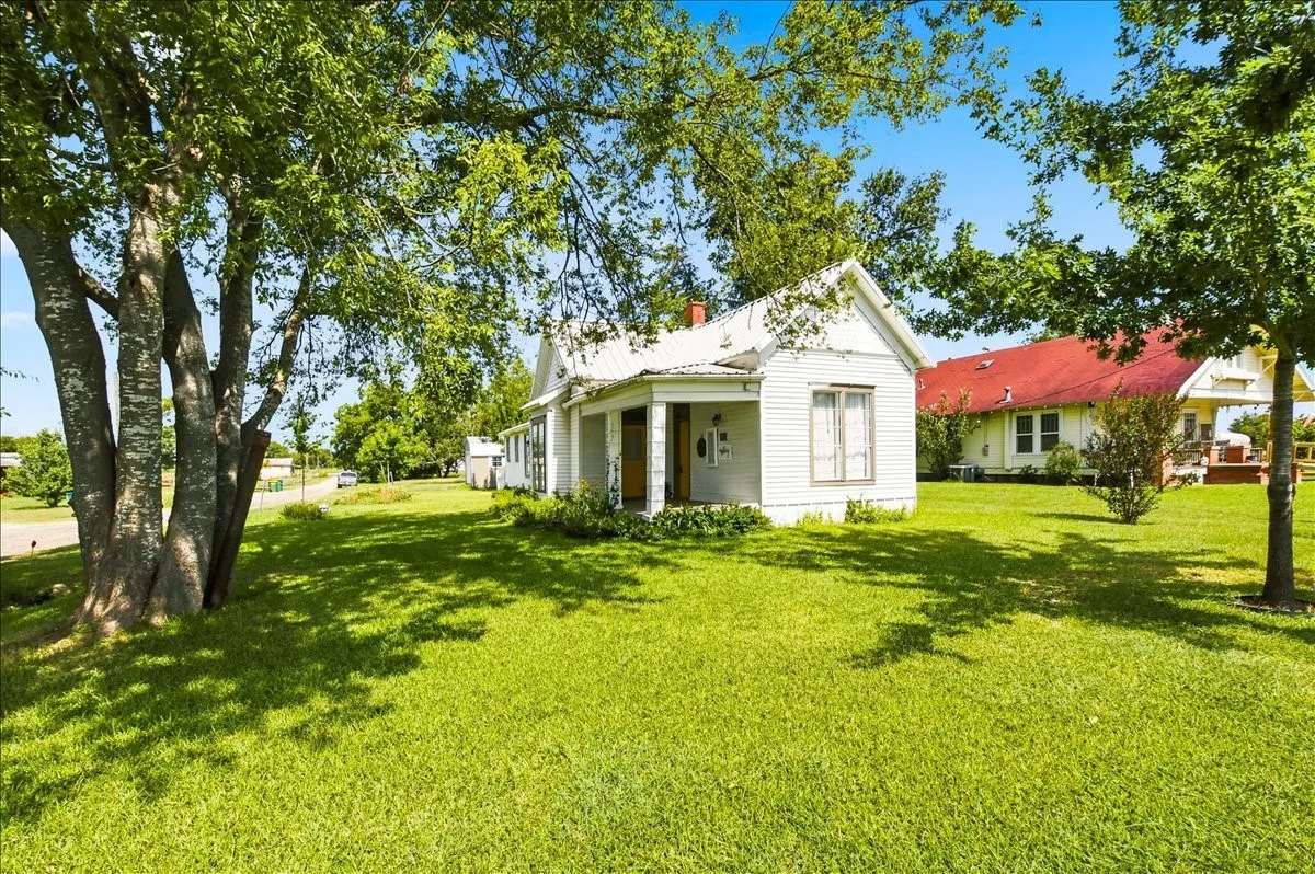 Angled front view of the white cottage home surrounded by mature trees and a vast lawn.