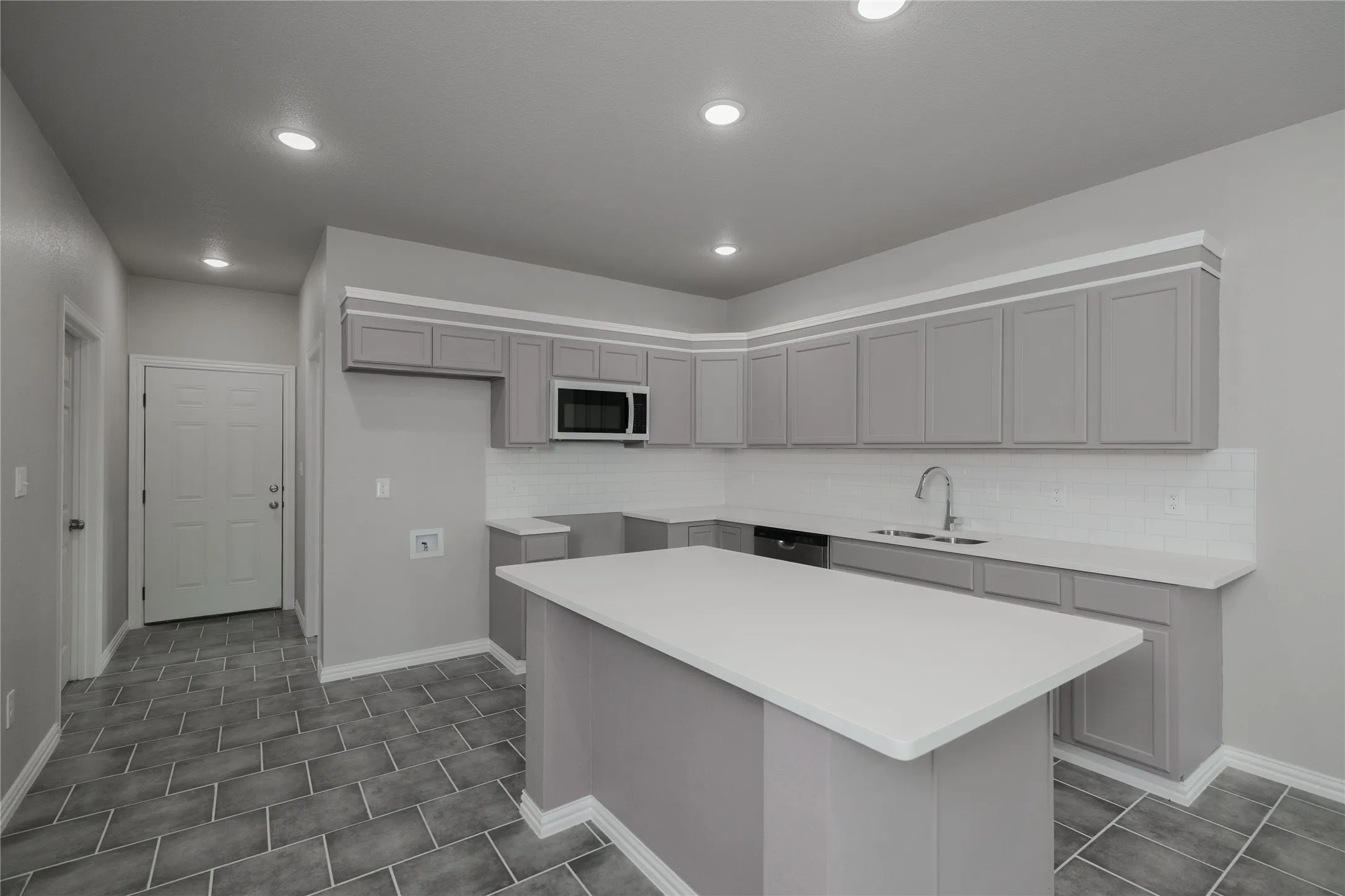 Modern kitchen featuring a central island with a white countertop, gray cabinetry, white subway tile backsplash, and stainless steel microwave