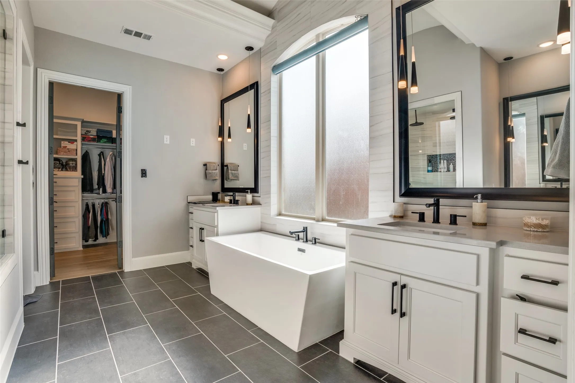 Spacious bathroom featuring a freestanding soaking tub, dual vanity with quartz countertops, custom cabinetry, and large format dark tile flooring