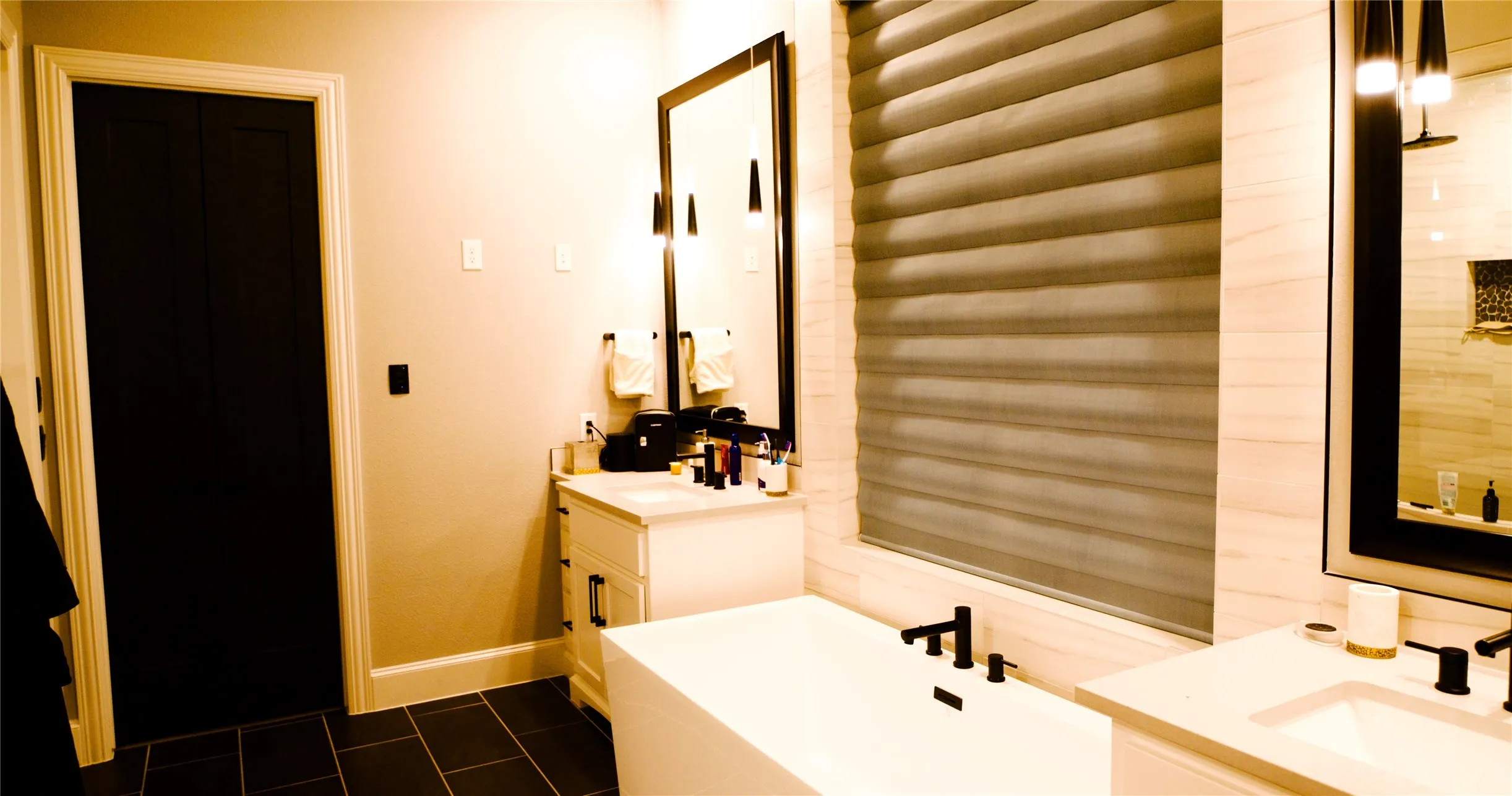 Modern bathroom featuring a freestanding tub with matte black fixtures, dual vanities with undermount sinks and quartz countertops, and a large window with horizontal blinds