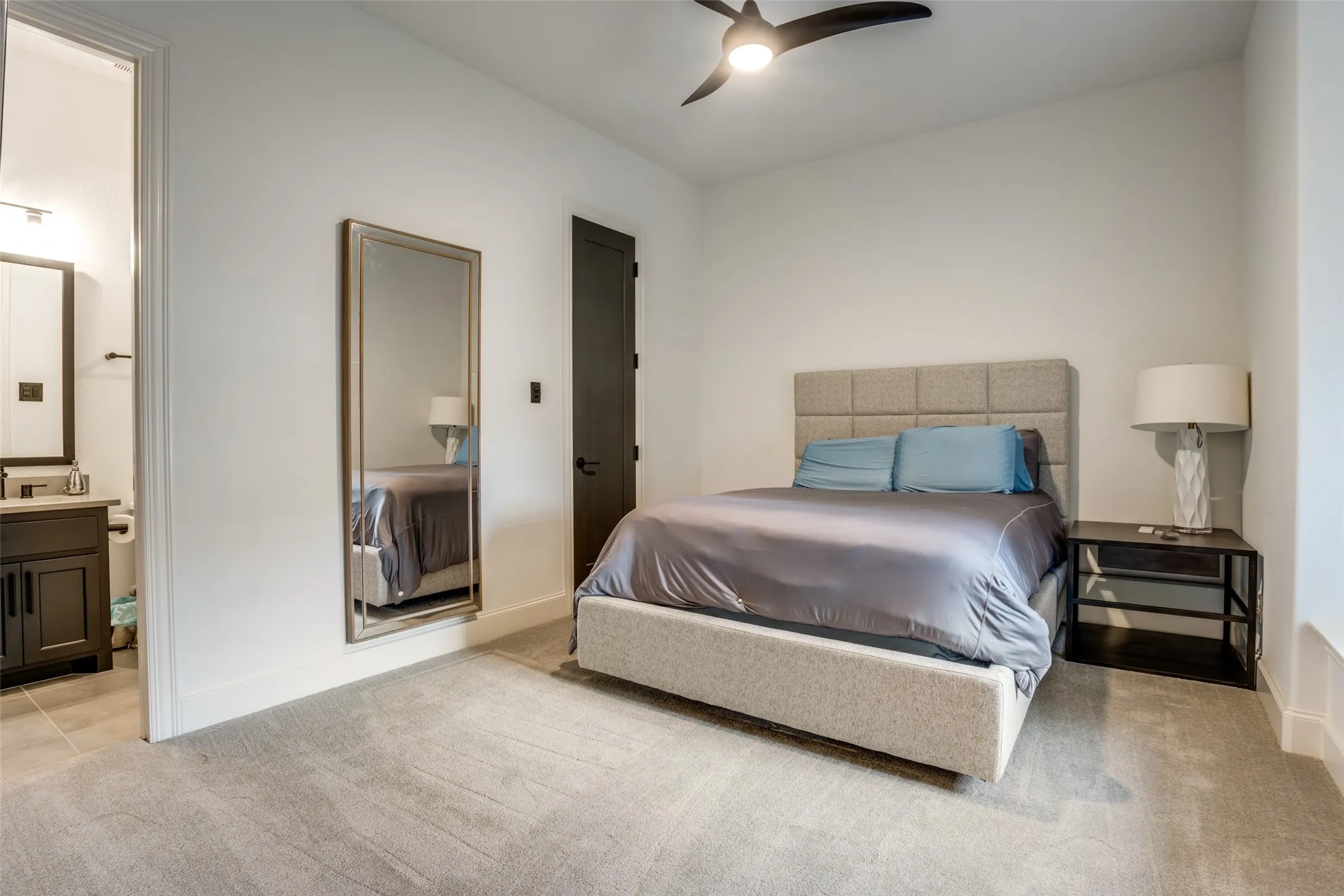 Carpeted bedroom featuring a dark-finish ceiling fan, a dark-finish door with black hardware, and an open doorway revealing a bathroom with a vanity and mirror