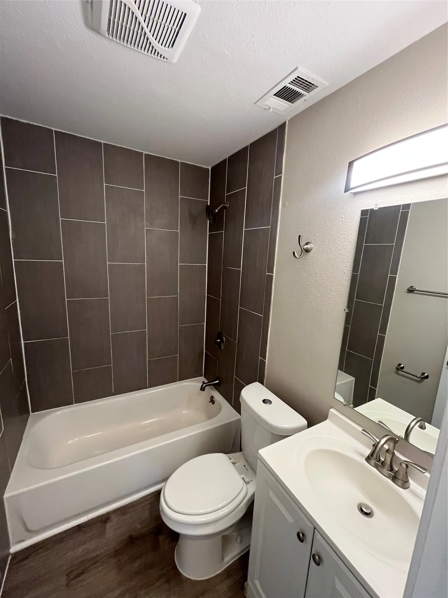 Contemporary bathroom featuring a white bathtub with dark tiled surround, a white toilet, a white vanity with integrated sink, and wood-finish flooring