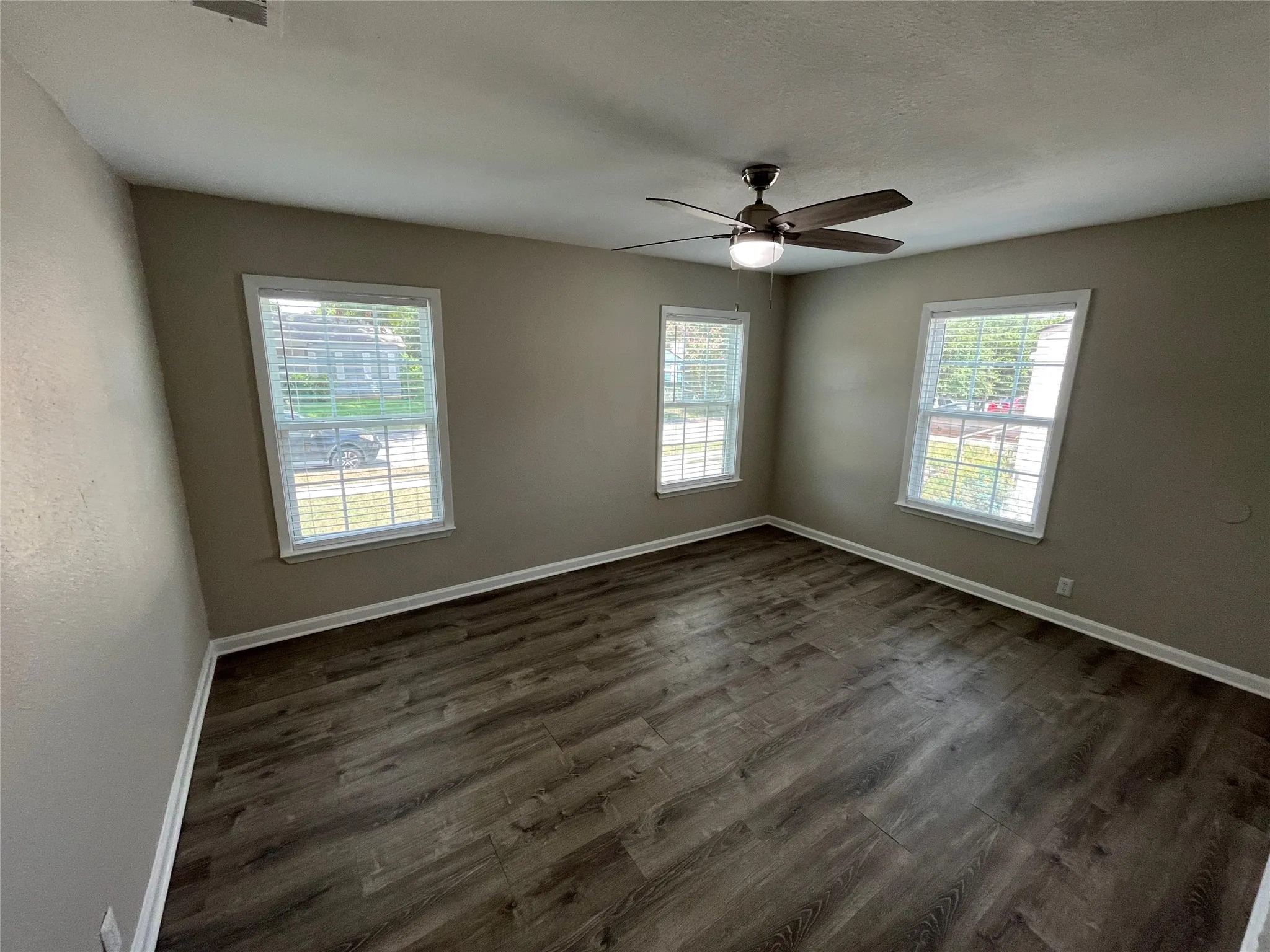Spacious room with wood-finish flooring, three windows with blinds, a ceiling fan with integrated lighting, neutral wall paint, and white trim
