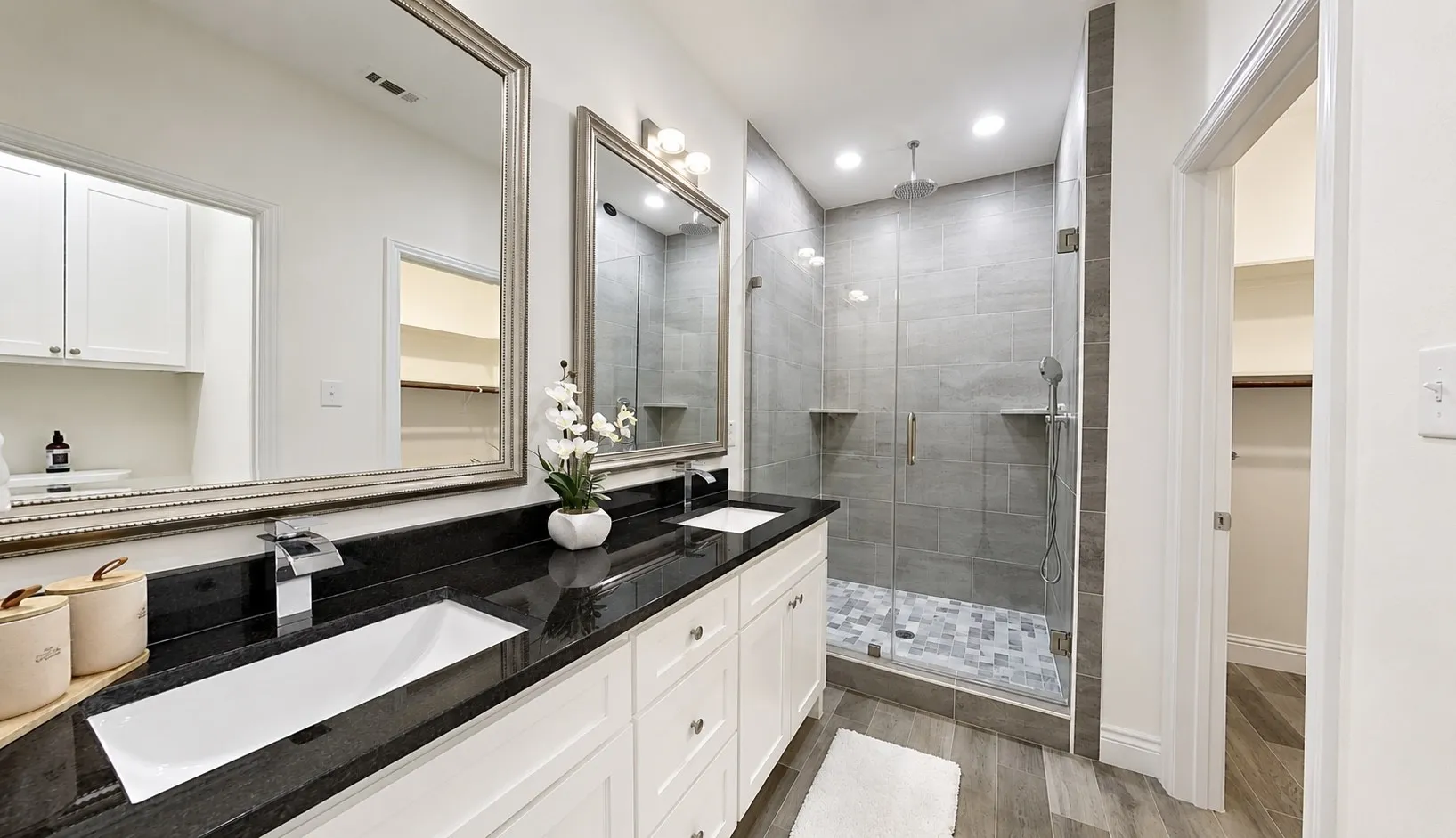Contemporary bathroom featuring dual rectangular undermount sinks set in a black countertop, white shaker-style vanity cabinetry, two framed wall mirrors, and a glass-enclosed shower with gray tiled walls and a mosaic tile​​‌​​​​‌​​‌‌​‌‌​​​‌‌​‌​‌​‌​​​‌​​ floor