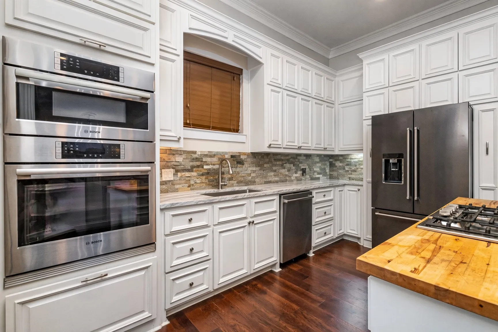 White cabinetry with crown molding, stainless steel built-in appliances, stacked stone backsplash, and wood-finish flooring