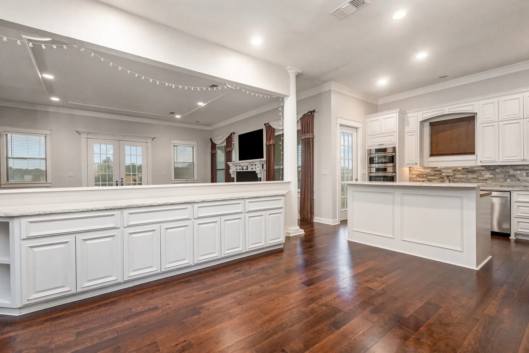 Wood-finish flooring throughout, white cabinetry, built-in double ovens, a central kitchen island, and a decorative tile backsplash