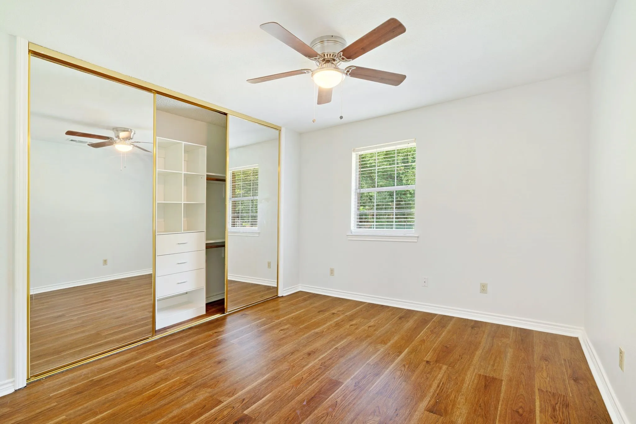 Spacious room featuring wood-finish flooring, a ceiling fan with light fixture, and a window with blinds