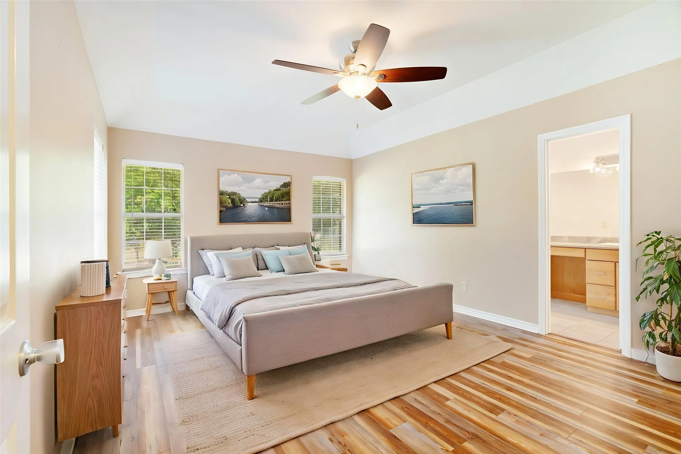 Vaulted ceiling, ceiling fan, wood-finish flooring, multiple windows with blinds, and an attached bathroom with a built-in vanity