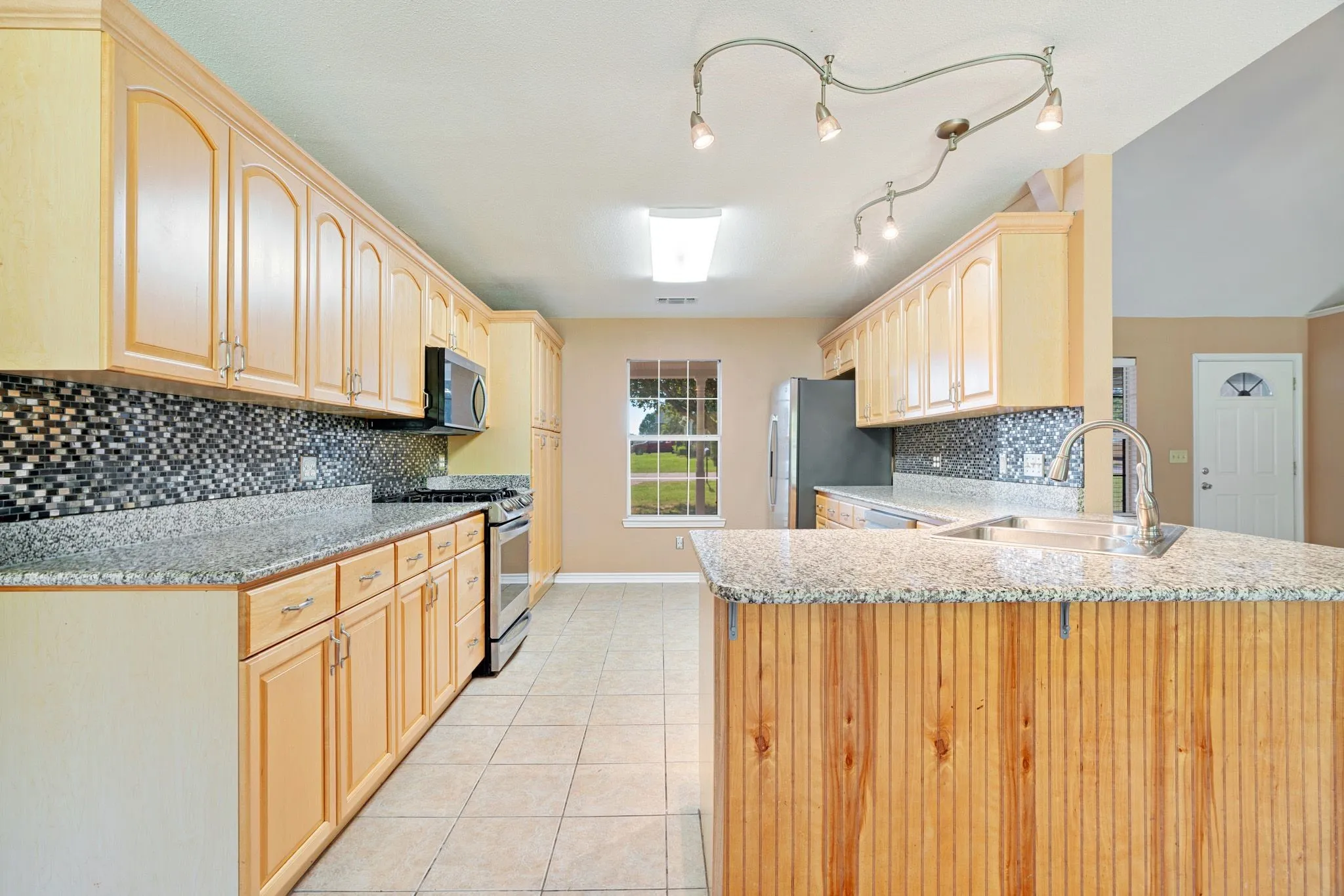 Open-concept kitchen featuring light wood cabinetry, stone countertops, a mosaic tile backsplash, stainless steel appliances, and a central island with an integrated sink