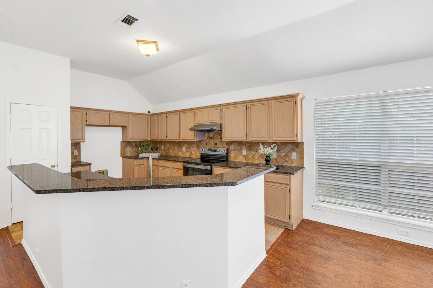 Kitchen featuring wood-finish flooring, a breakfast bar with stone countertops, wood cabinetry, a tiled backsplash, and a large window with horizontal blinds