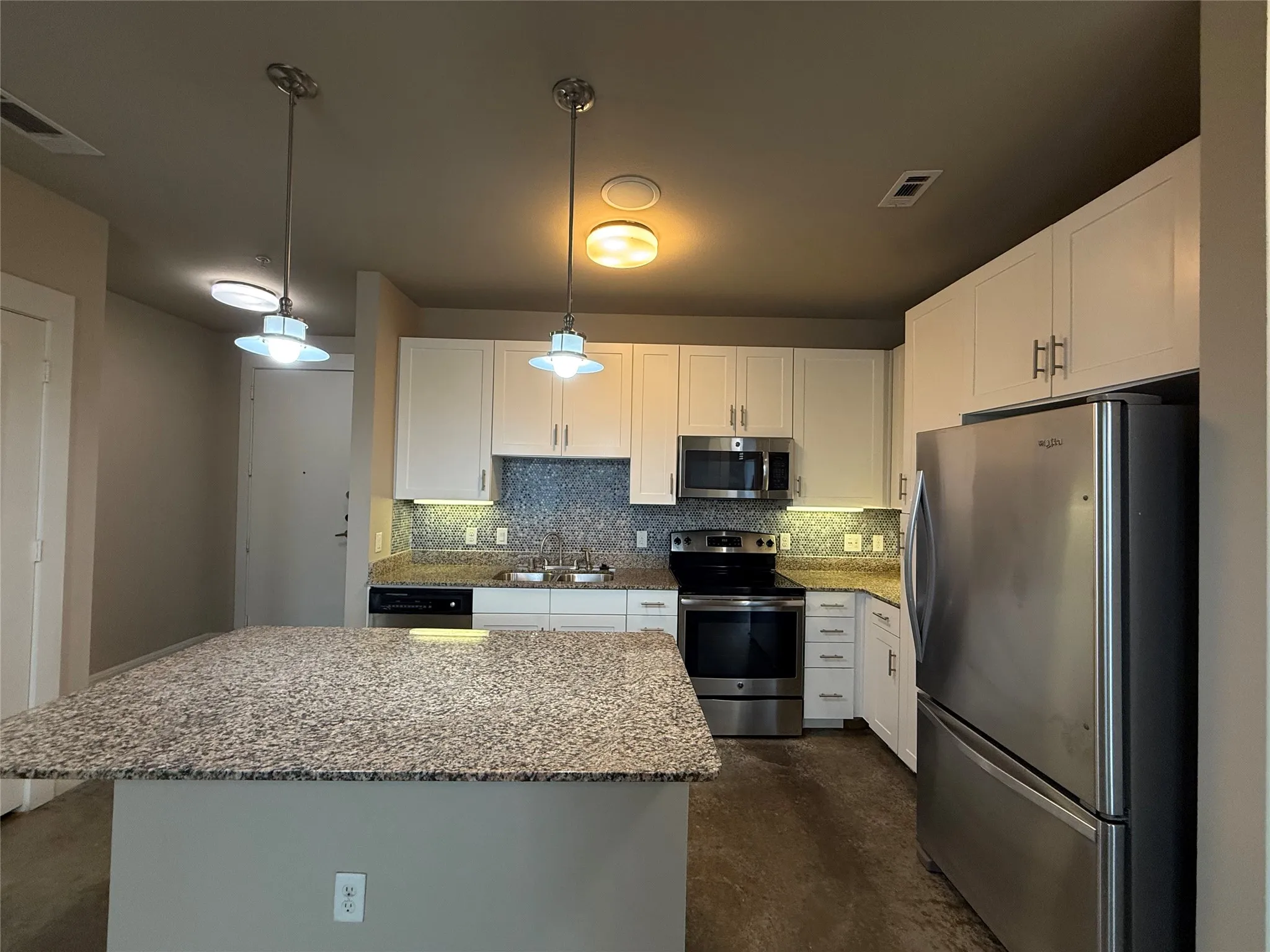 Kitchen featuring a central island with a speckled countertop, white cabinetry, stainless steel appliances, a mosaic tile backsplash, and two pendant lights