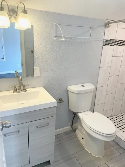 Bathroom featuring a single vanity with a white countertop and light wood-finish cabinetry, a commode, a shower with white and black mosaic tile flooring, and gray rectangular wall tile with a decorative accent band