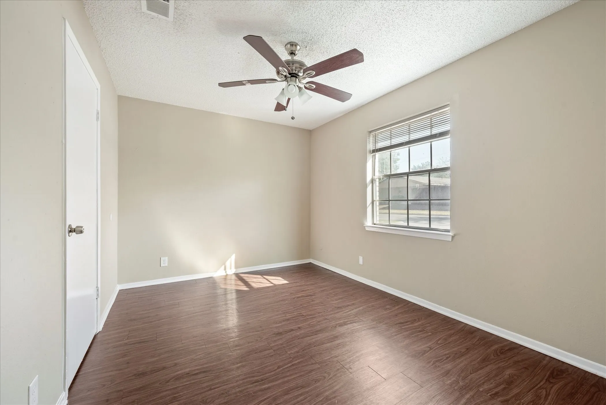 Spacious room featuring wood-finish flooring, neutral-toned walls, a ceiling fan with integrated lighting, and a multi-pane window