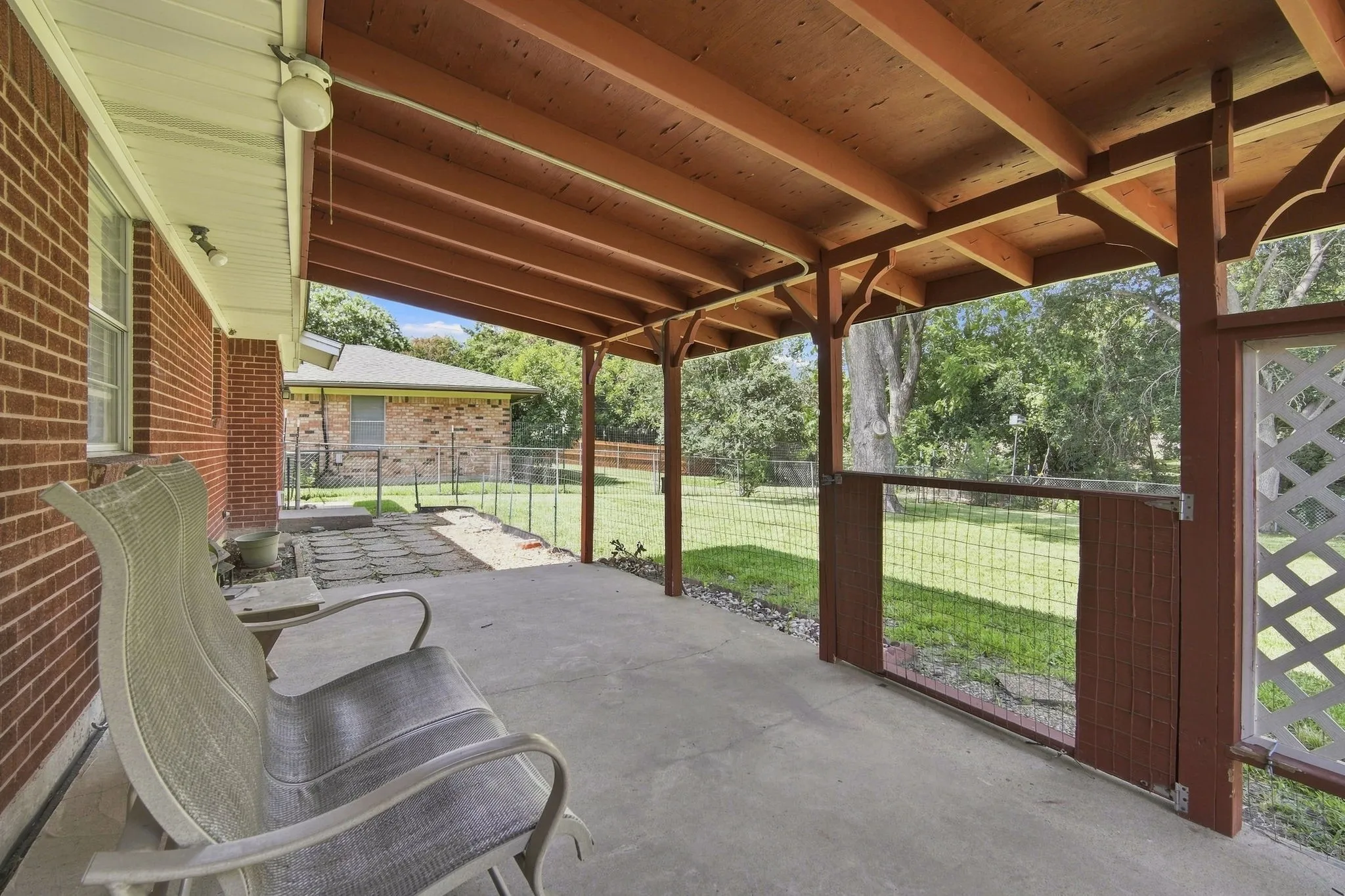 Covered patio featuring a concrete slab, exposed wood beam ceiling, brick exterior, lattice privacy screen, and wire fencing