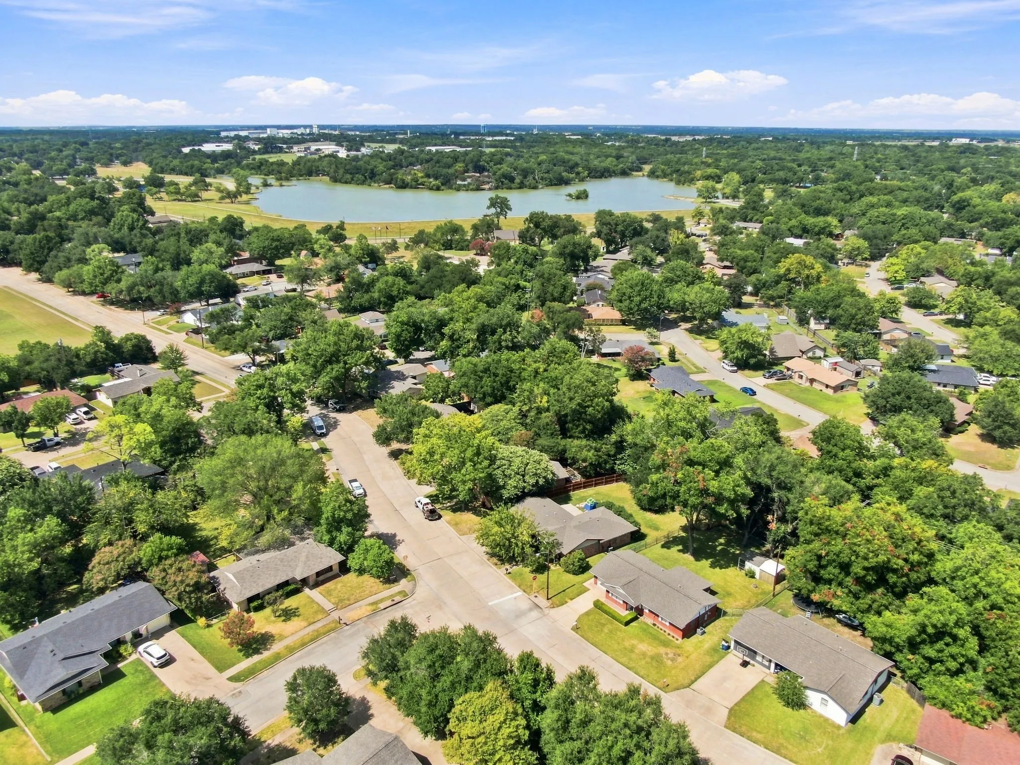 Aerial perspective showcasing a residential neighborhood with mature tree coverage, a central paved street, and a prominent body of water in the background