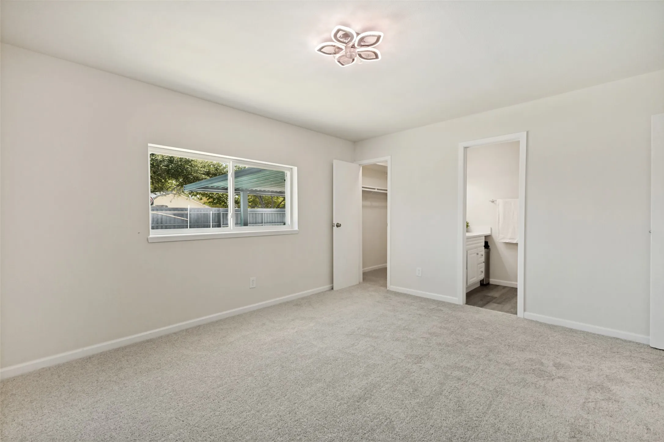 Light gray carpeting and painted walls with a window, ceiling light fixture, open closet, and an entry to an additional room featuring a white vanity