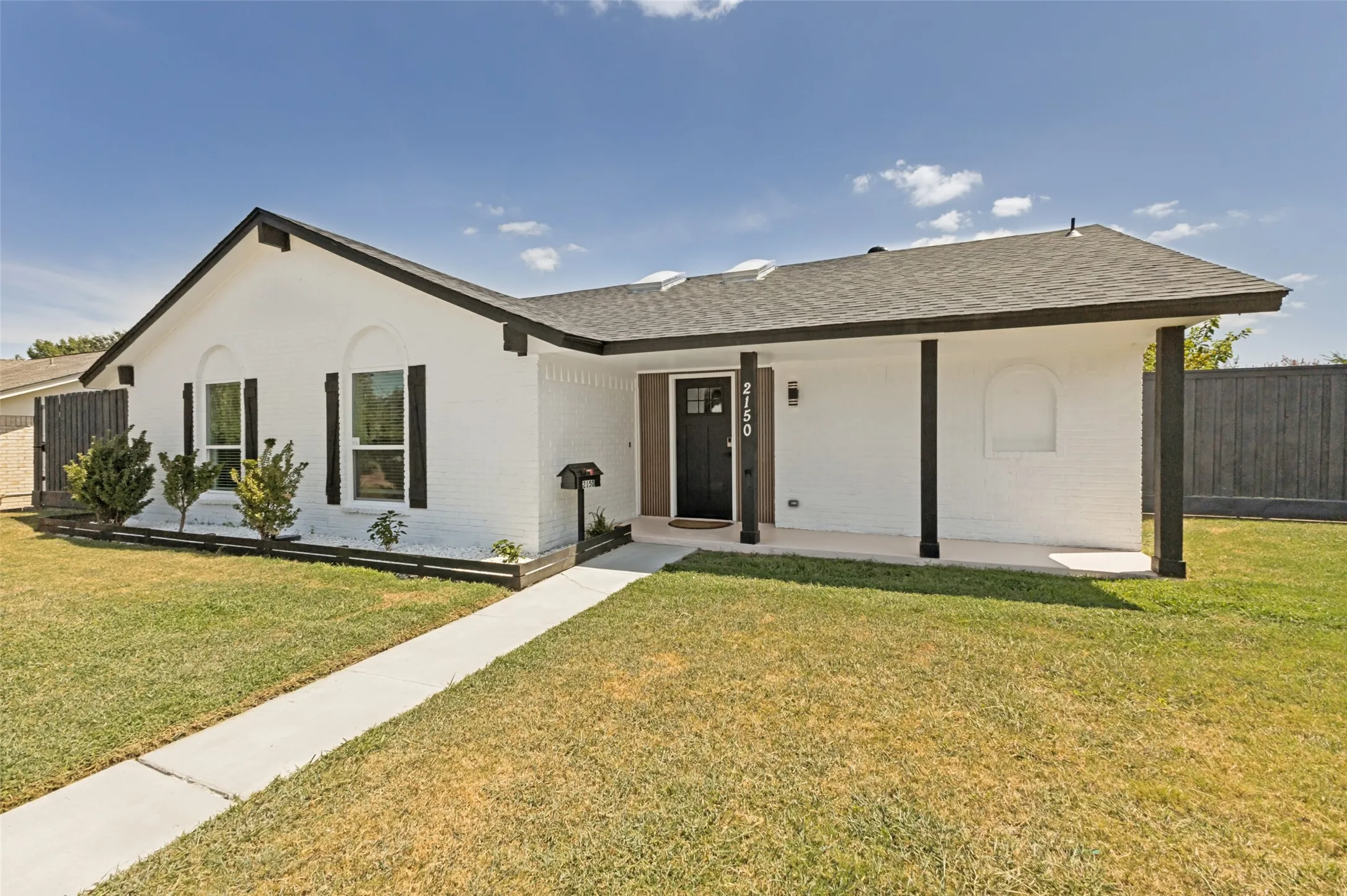 White brick exterior featuring a covered entry, dark trim accents, a concrete walkway, and a landscaped front yard with a wooden fence