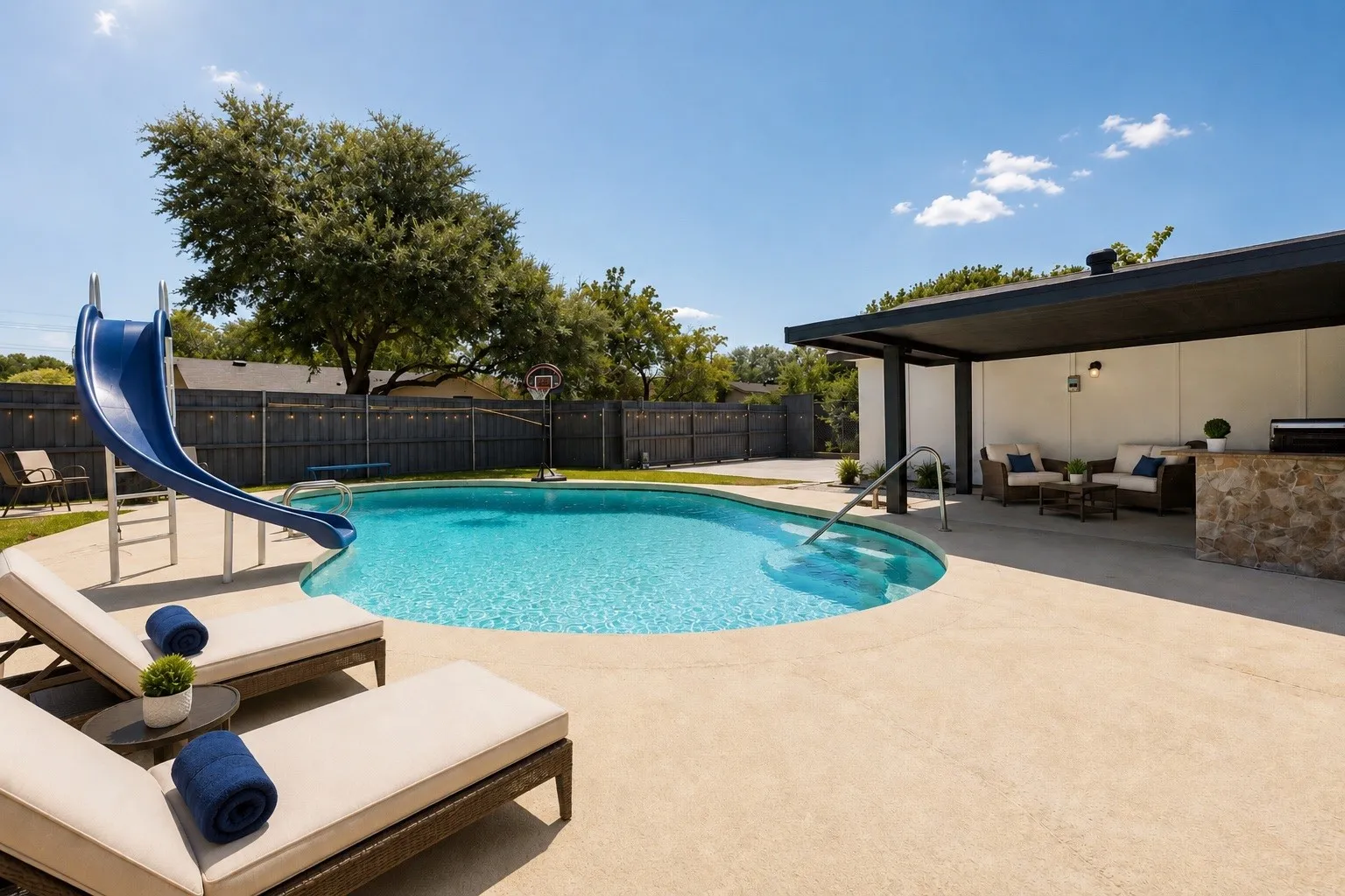 Private backyard oasis featuring a freeform swimming pool with a slide, a built-in outdoor kitchen with stone detailing, an expansive concrete patio, and mature perimeter trees
