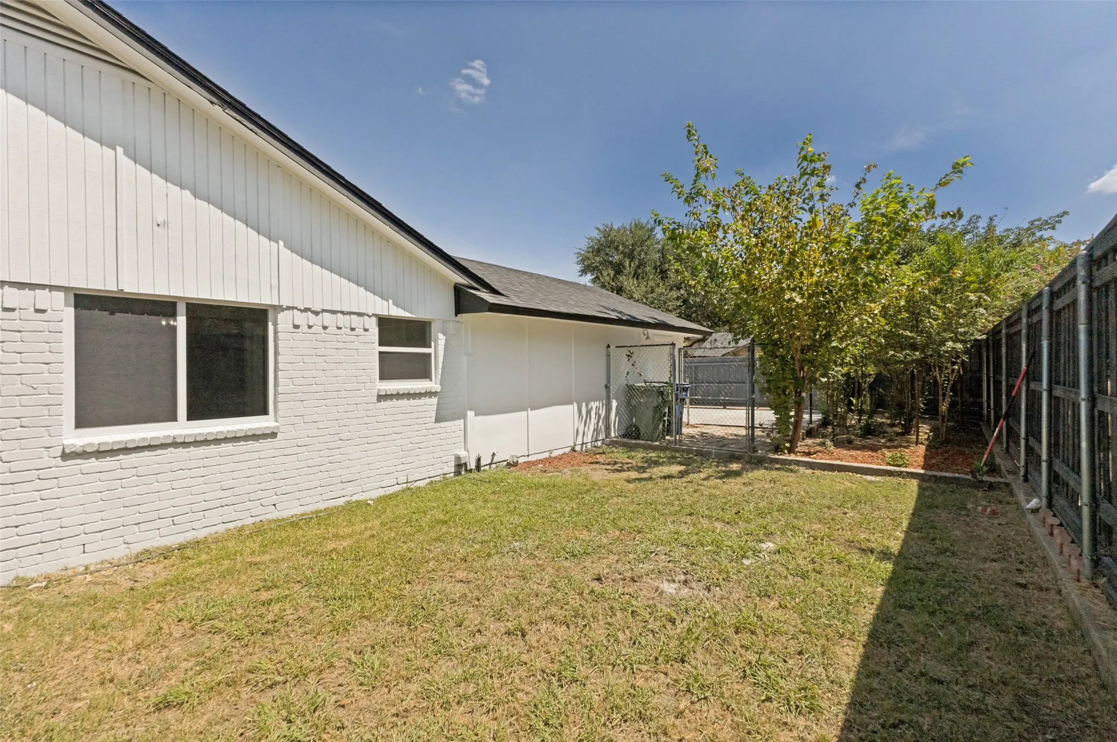 Side yard featuring a white painted brick and vertical siding exterior, two windows with screens, a dark shingle roof, established trees, and a dark privacy fence