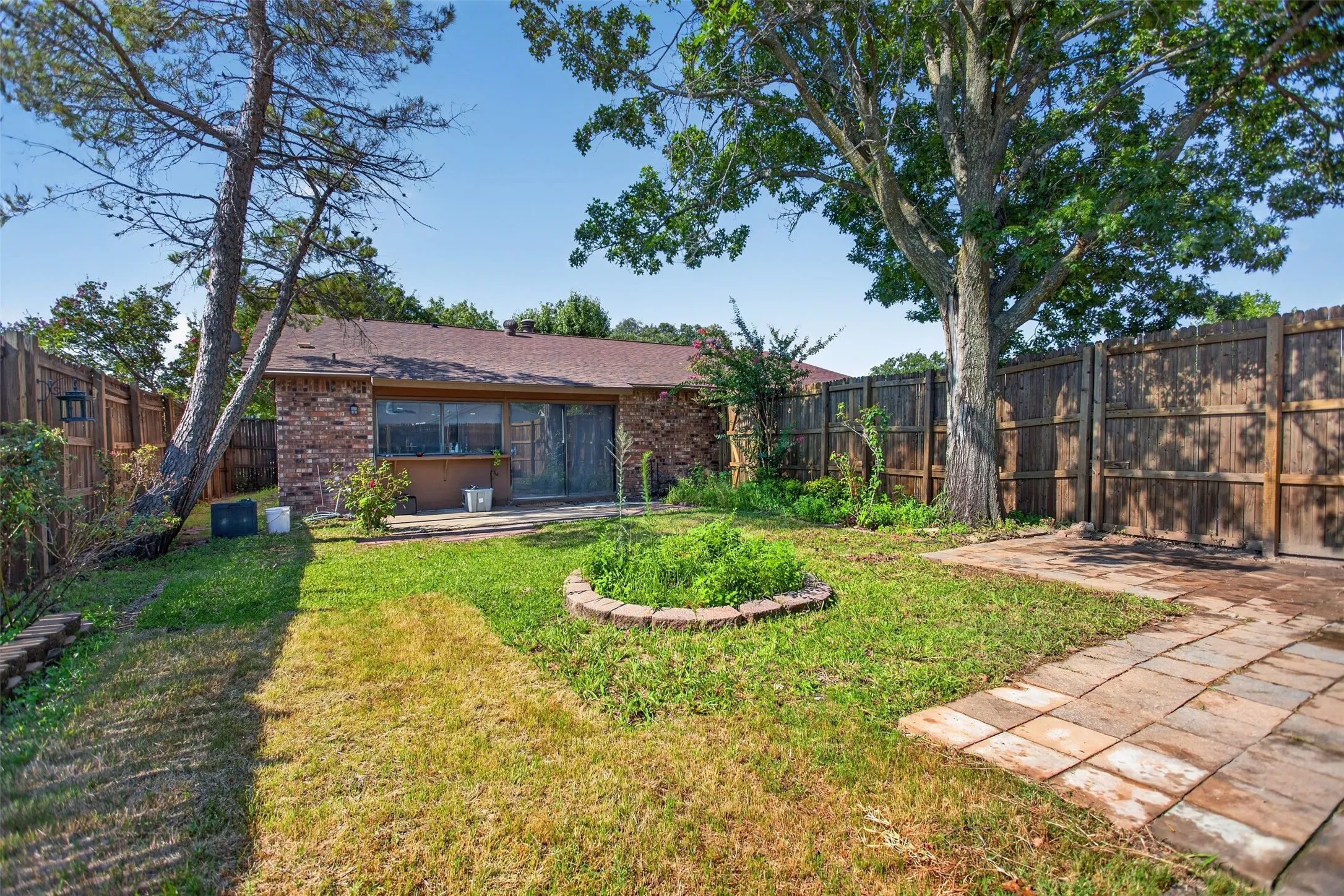 Expansive fenced backyard featuring a brick exterior, a mature shade tree, a stone paver patio, and a central planting bed