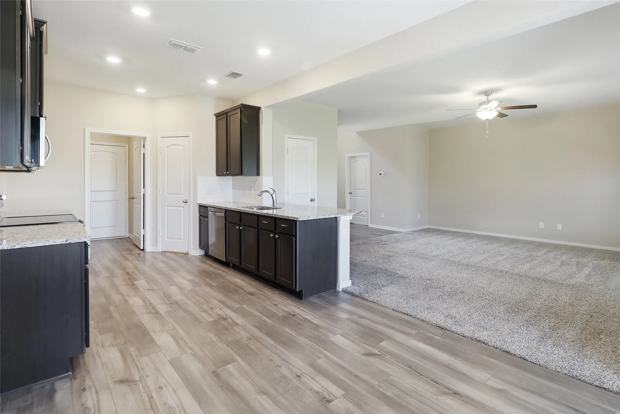 Open-concept living area featuring wood-finish flooring, dark wood cabinetry, light countertops, and a ceiling fan in the carpeted portion