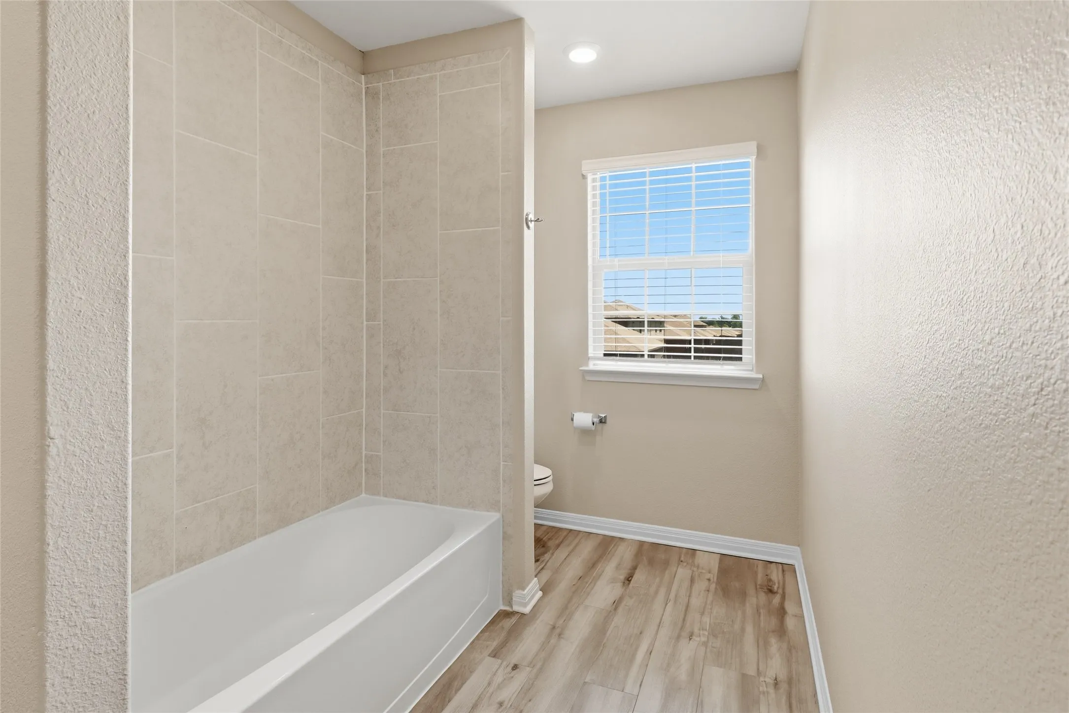 Bathroom featuring a tub/shower combination with light-toned tiled surround, wood-finish flooring, a window with blinds, and recessed lighting