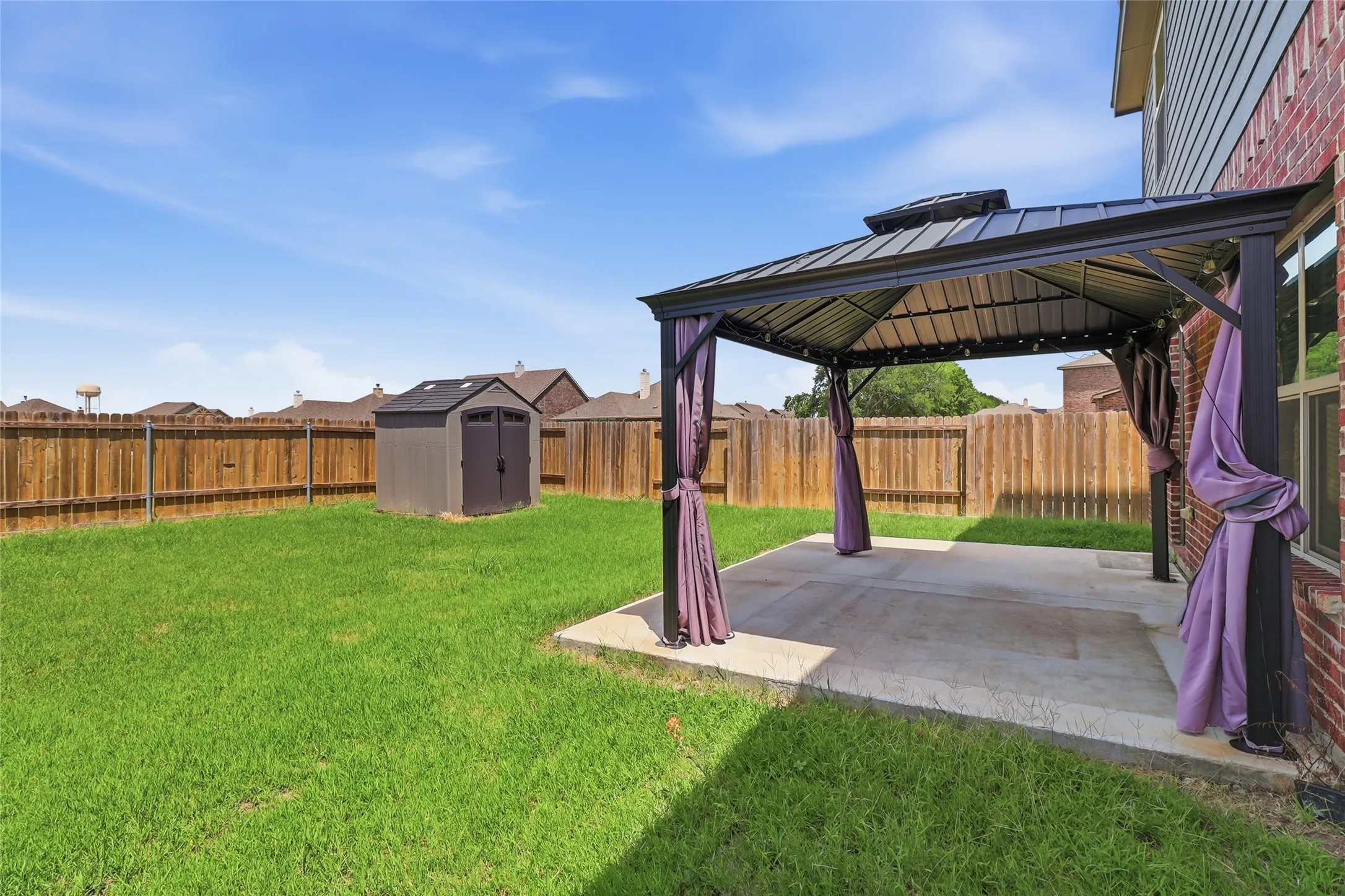 Expansive backyard featuring a concrete patio with a permanent gazebo, a detached storage shed, lush green lawn, and a wood privacy fence