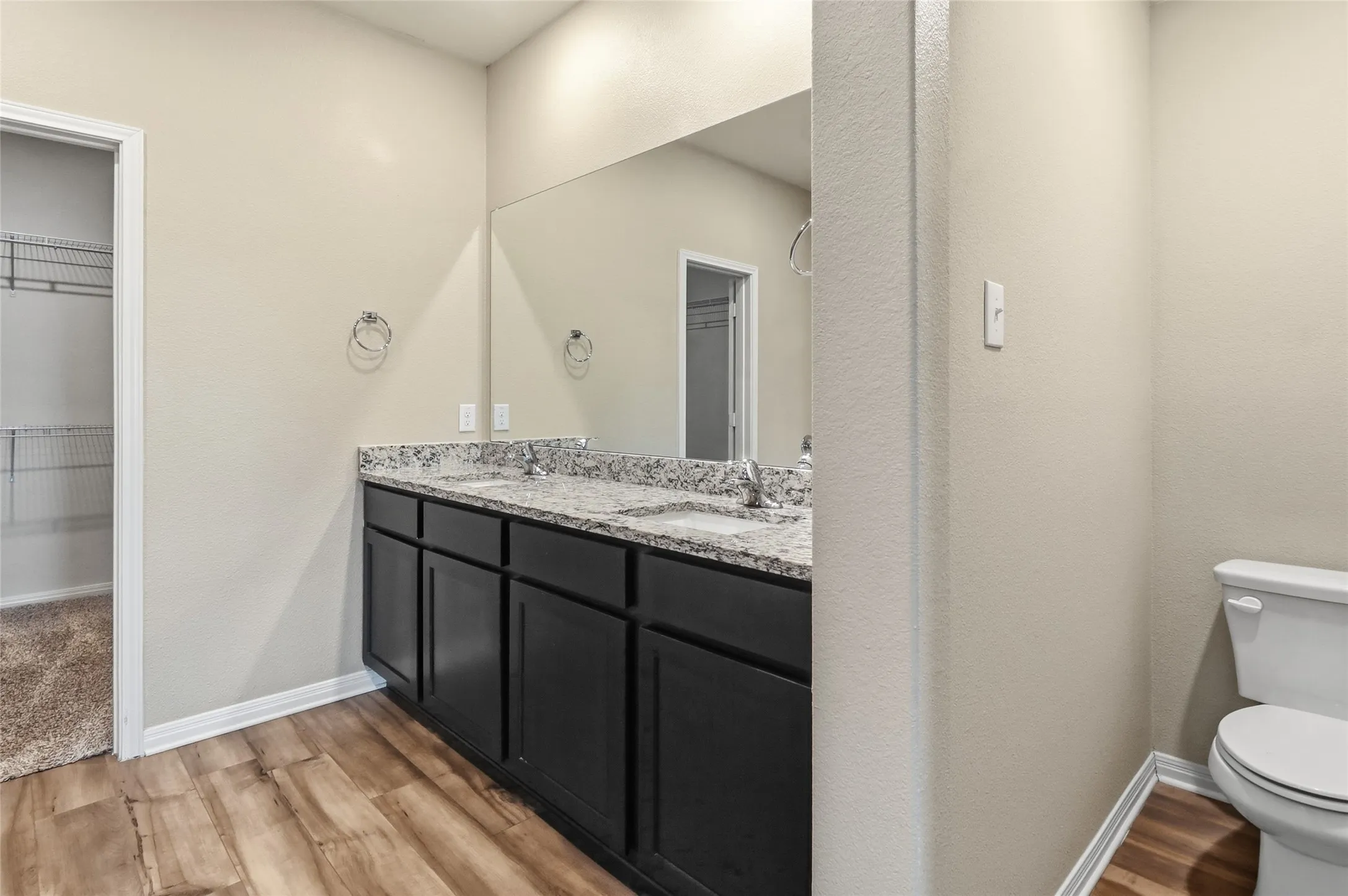 Secondary Bathroom featuring a dual vanity with a granite-look countertop and dark cabinetry, wood-finish flooring, and a full-width mirror