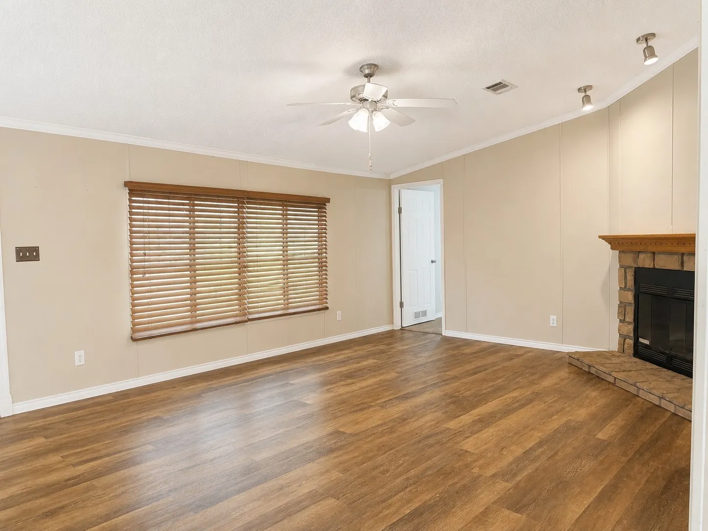 Spacious living area featuring wood-finish flooring, a ceiling fan, and a corner fireplace with a stone surround and wood mantel